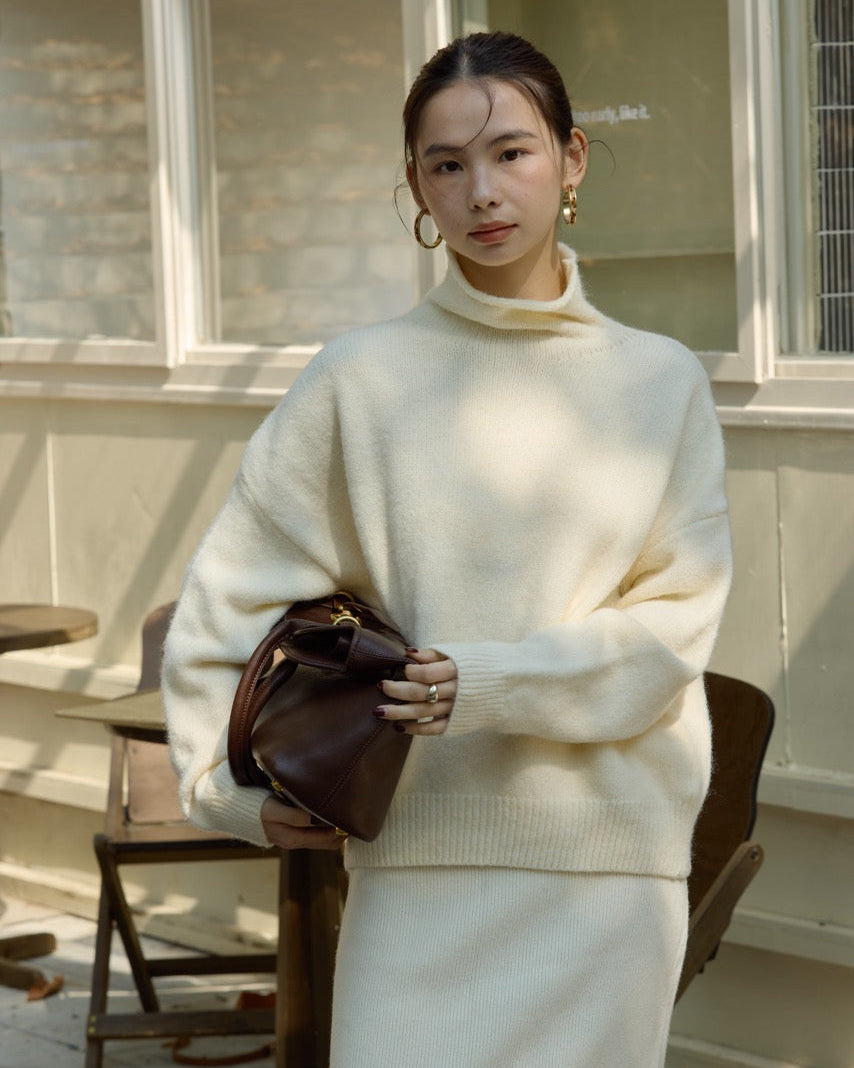 Woman wearing a cream sweater and skirt holding a brown handbag indoors.