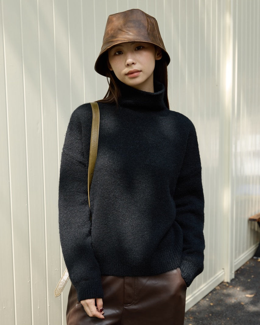 woman wearing a black turtleneck sweater and brown bucket hat standing against a light-colored wall.
