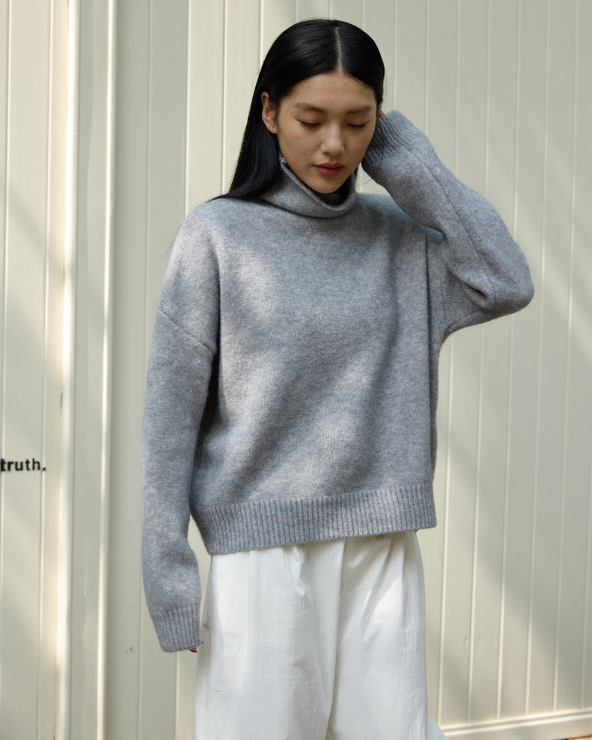 Person wearing a gray sweater and white pants against a light-colored wall.