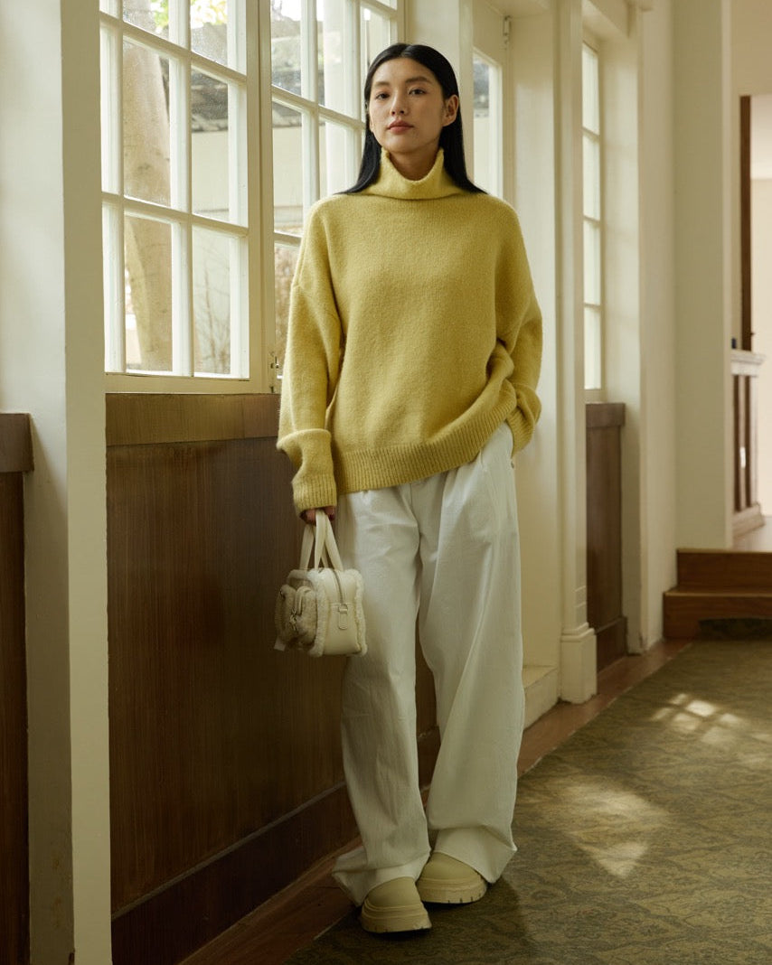 Woman in a yellow sweater and white pants standing in a sunlit room.
