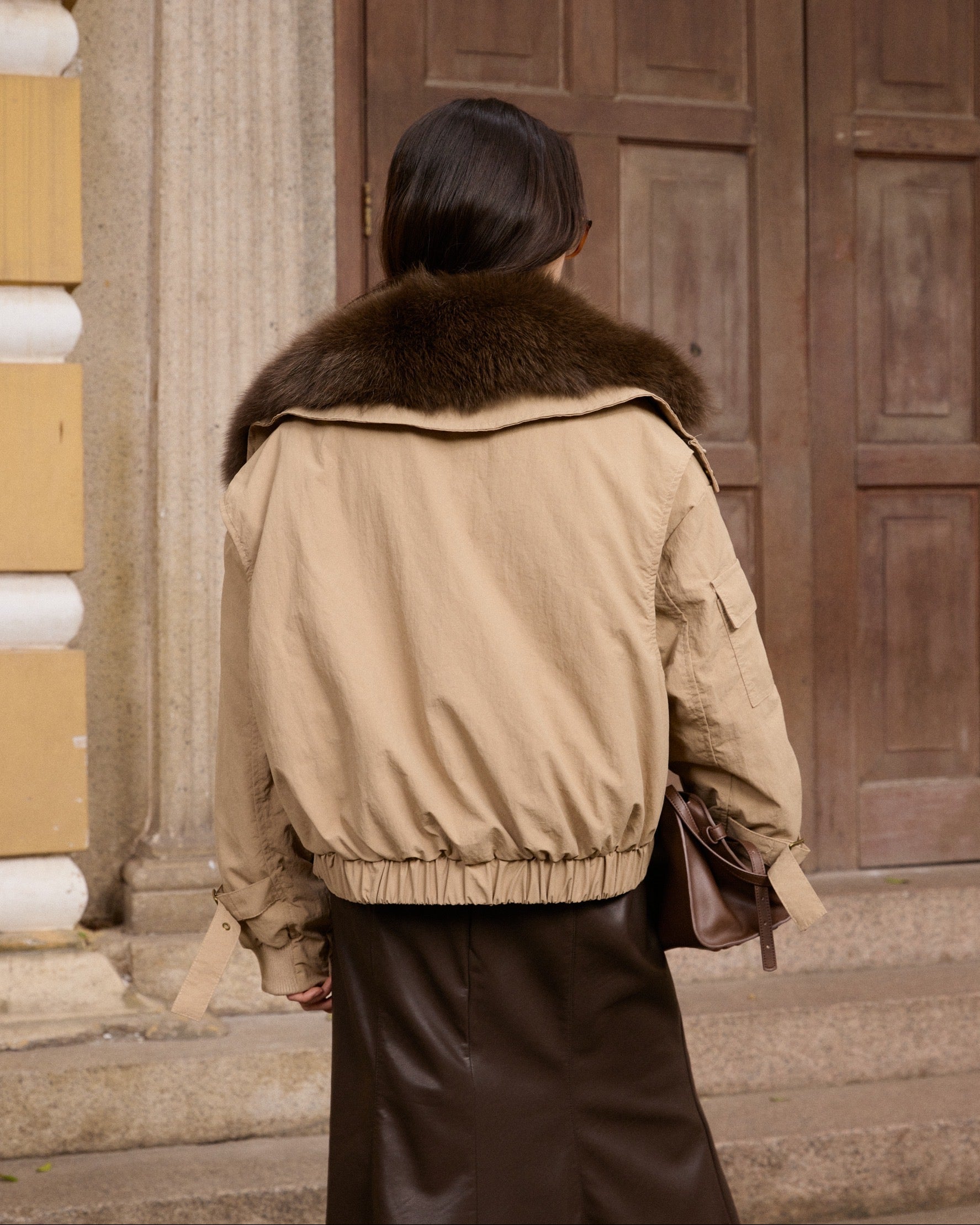 woman wearing a beige bomber jacket with fur collar in front of a wooden door.