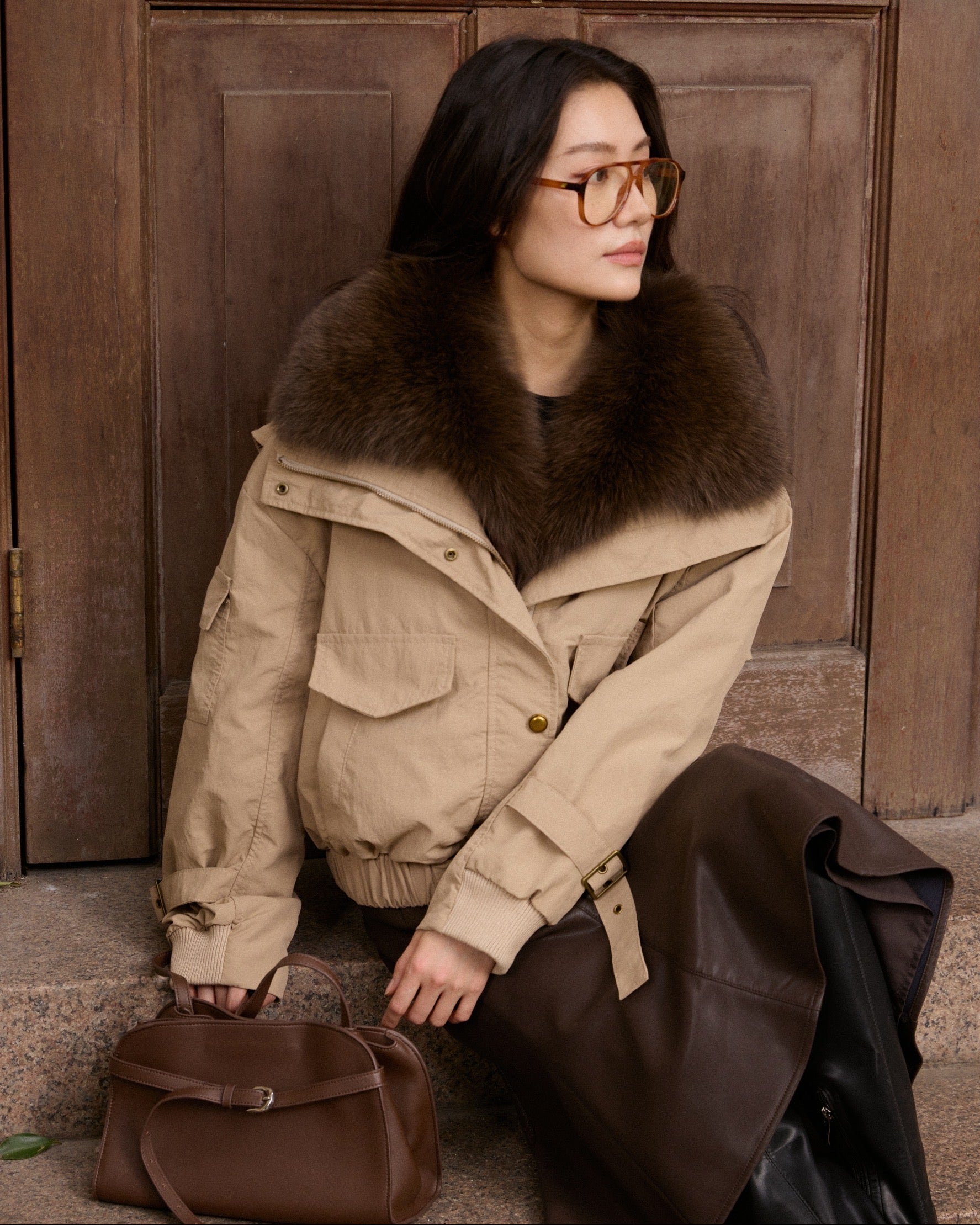 Woman wearing a beige coat with a fur collar, sitting against a wooden door.