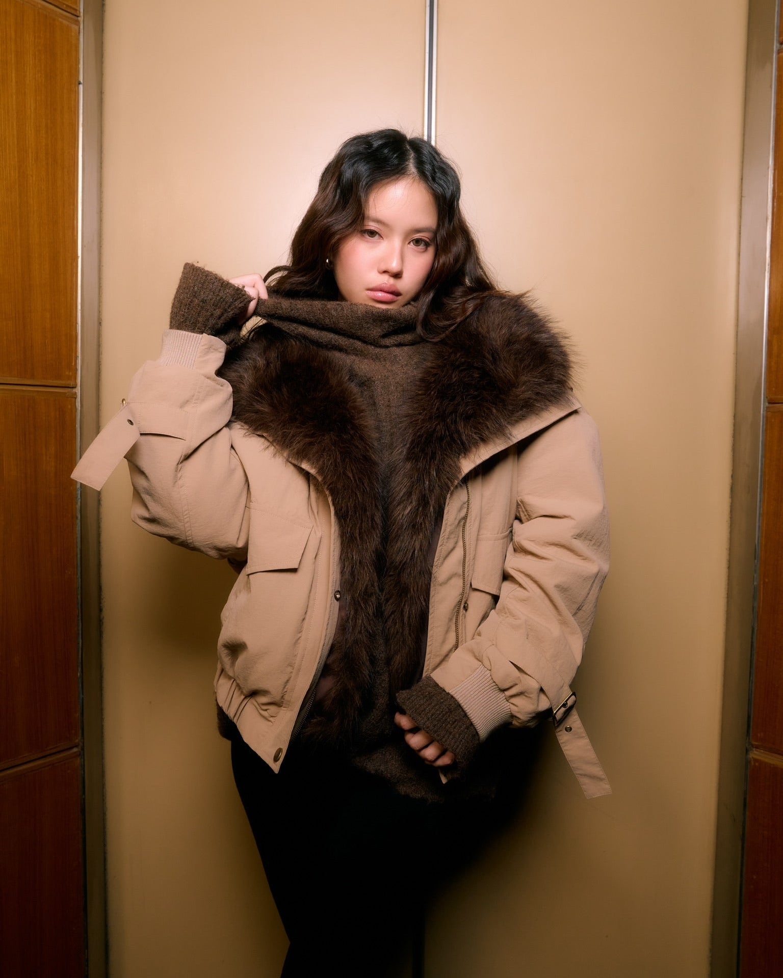 woman wearing a large beige coat with fur trim in an elevator.