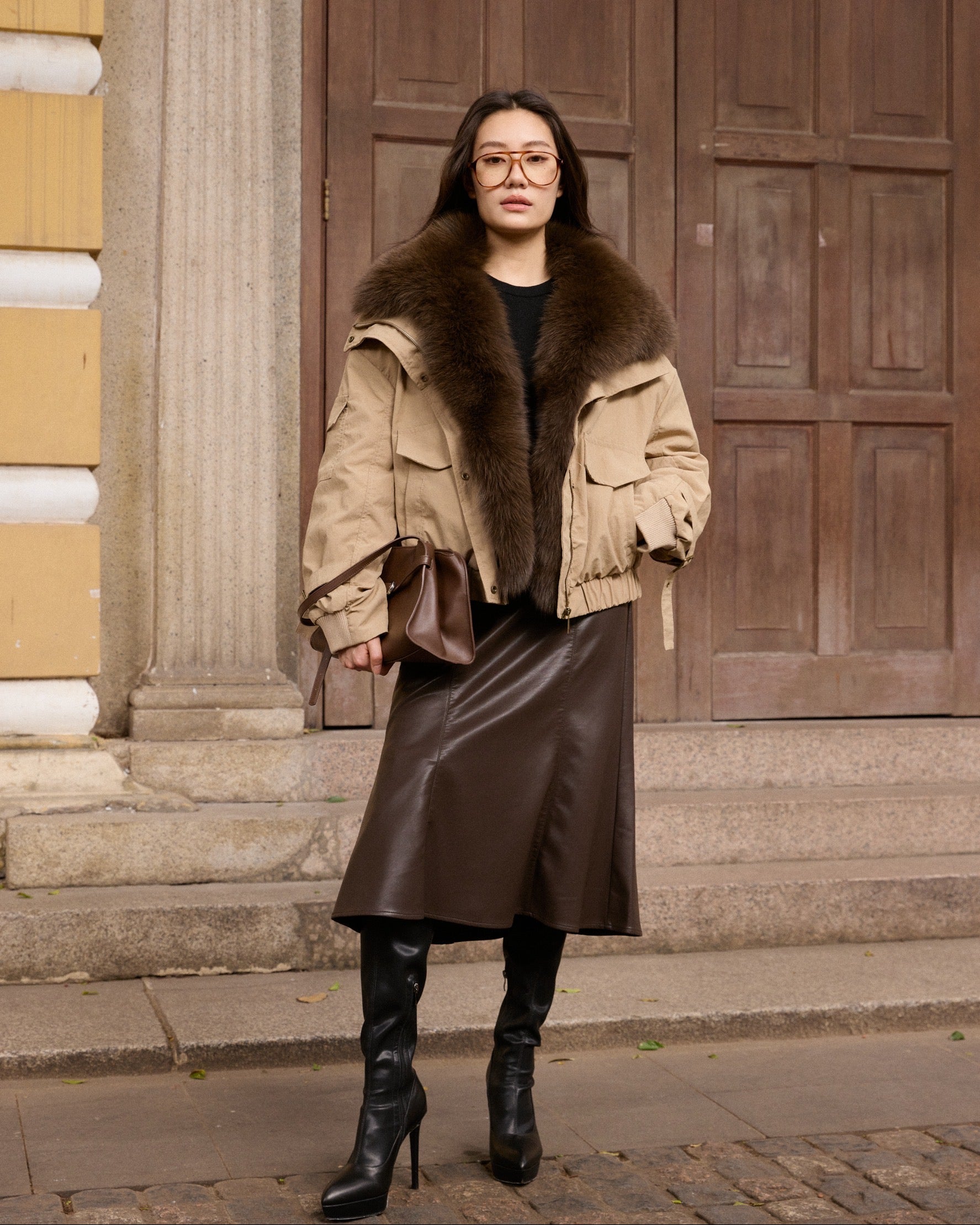 Woman wearing a fur-trimmed coat and leather skirt standing in front of a building.