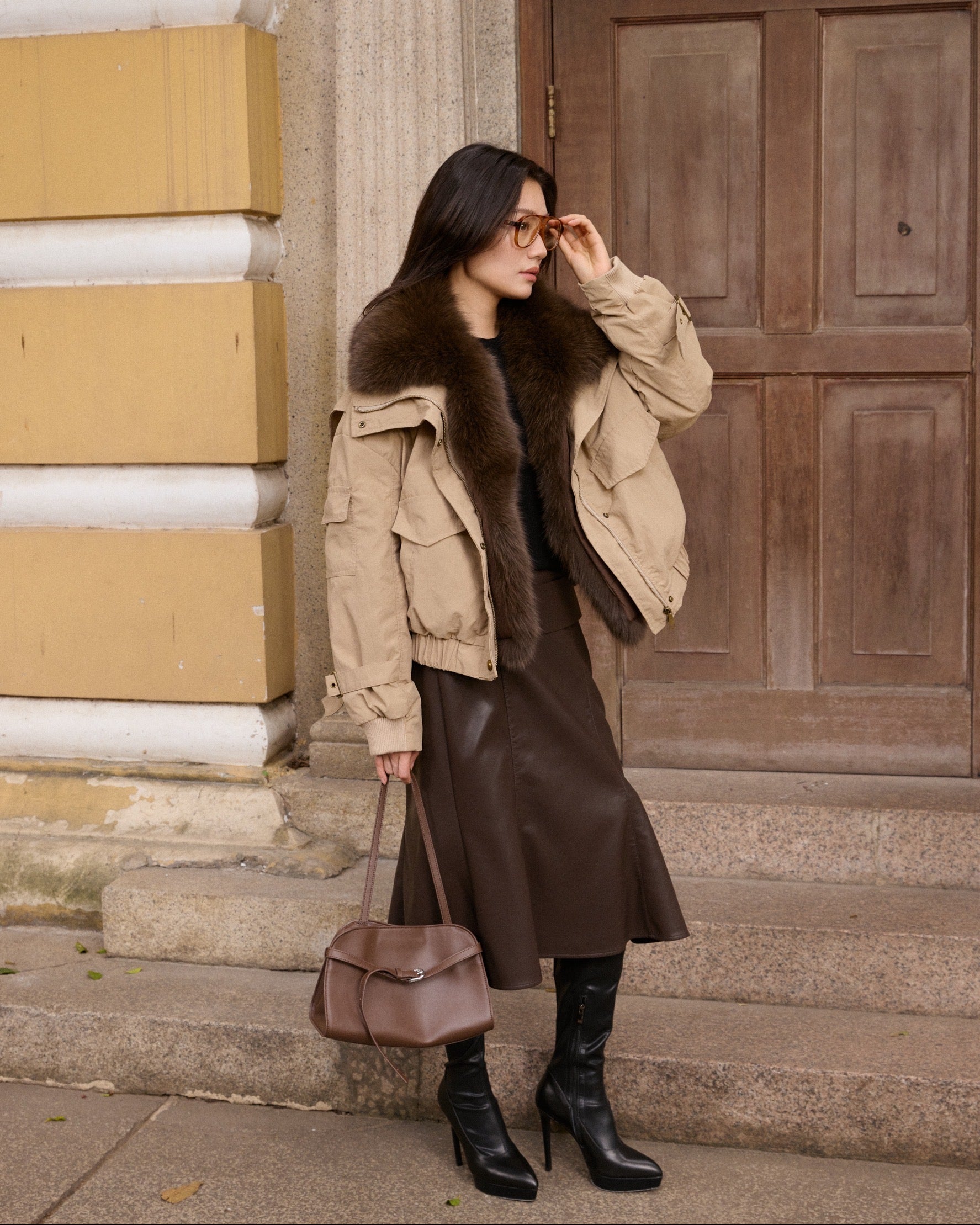 Woman in a beige coat with fur collar standing on steps in front of a building.