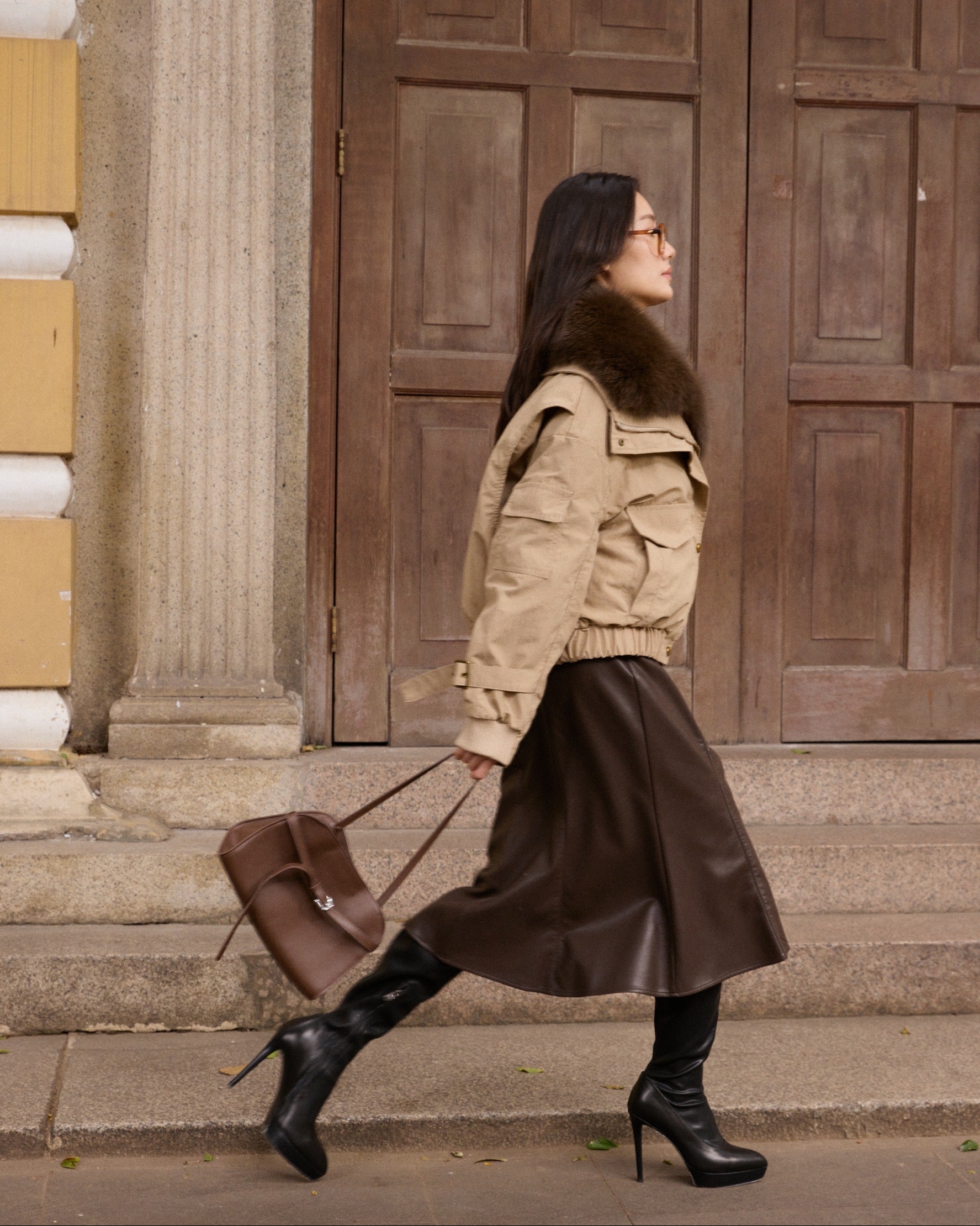 Woman in beige jacket and brown leather skirt walking past a large wooden door.