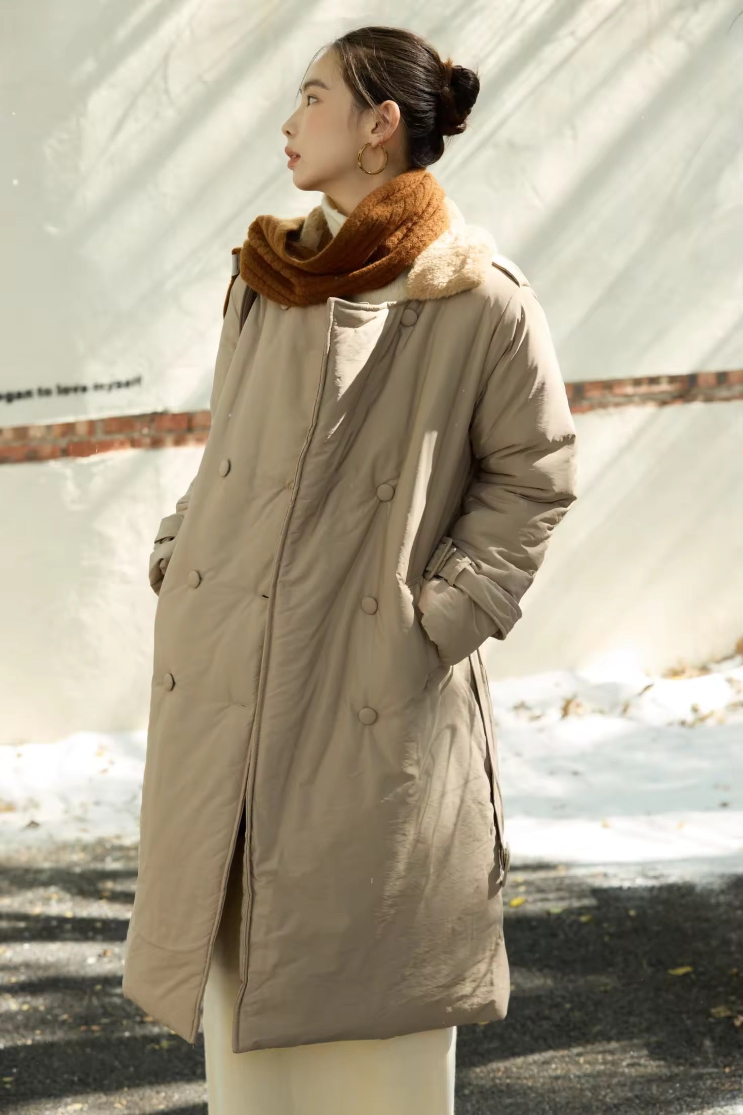 Woman wearing a beige coat and brown scarf standing outdoors.