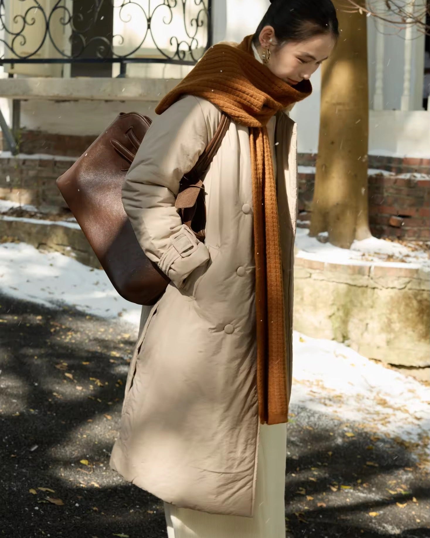 woman wearing a beige coat and brown scarf in an outdoor setting with snow on the ground.