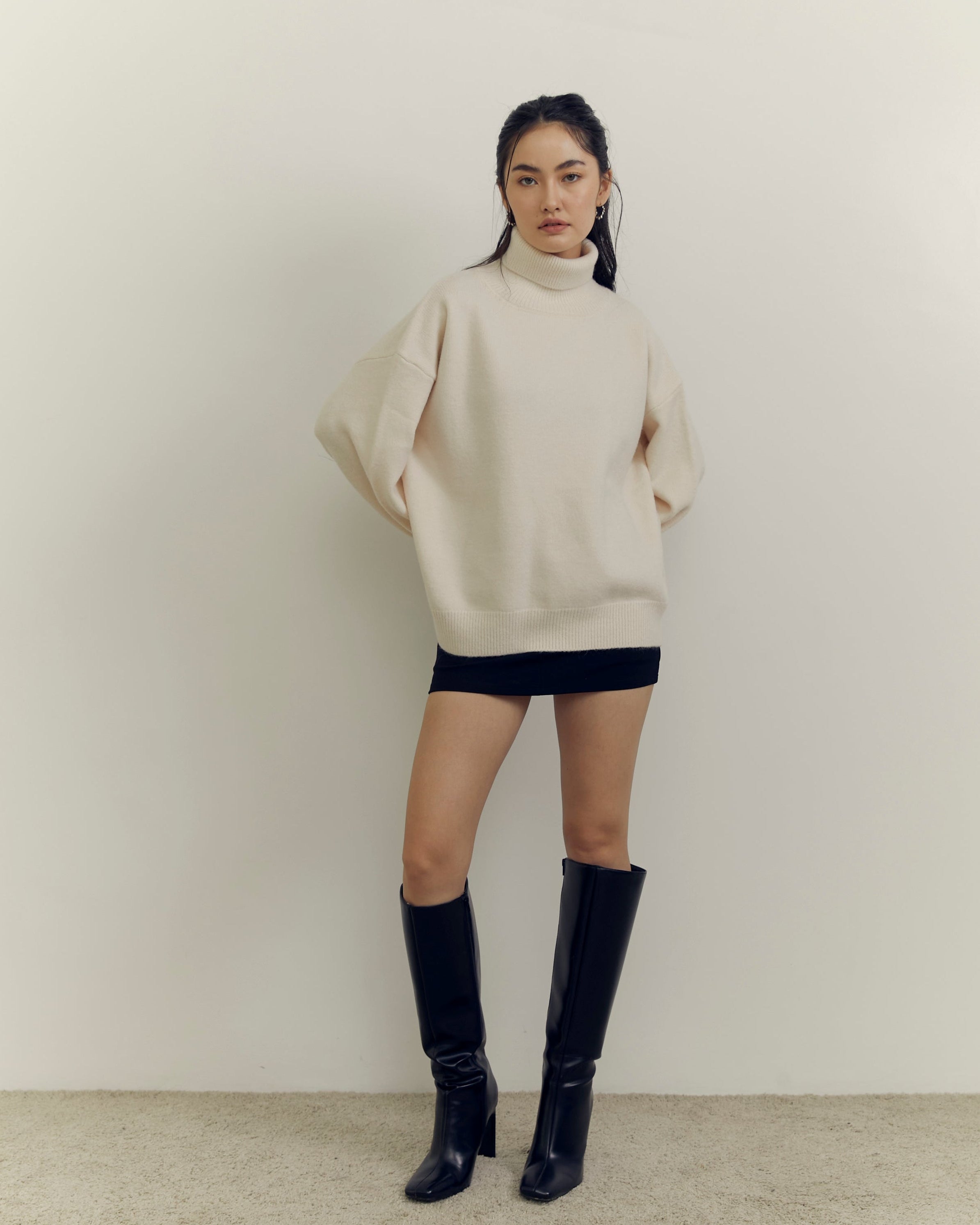 Person wearing a beige sweater and black knee-high boots against a plain background