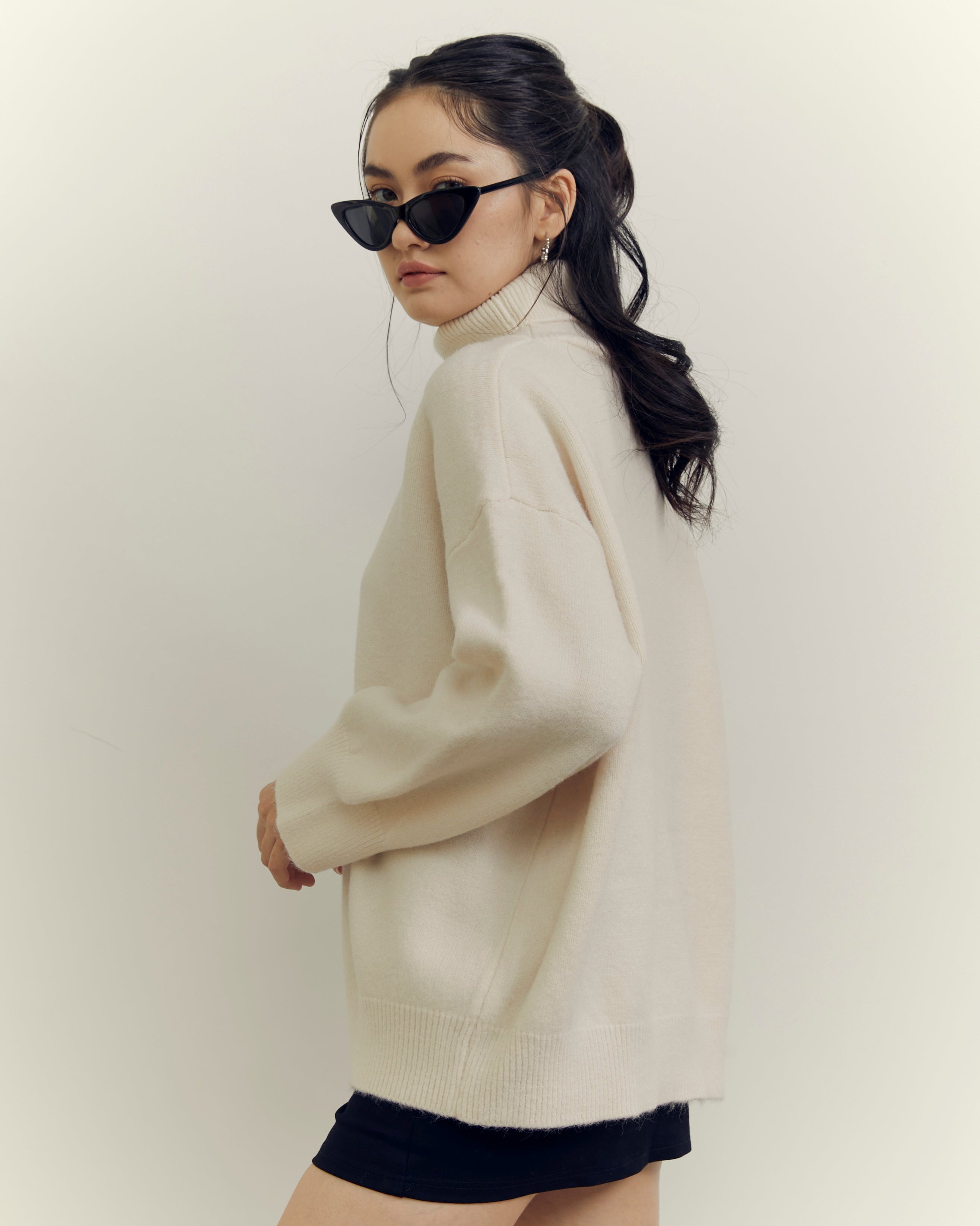 Woman wearing a beige sweater and black shorts with sunglasses on a plain background