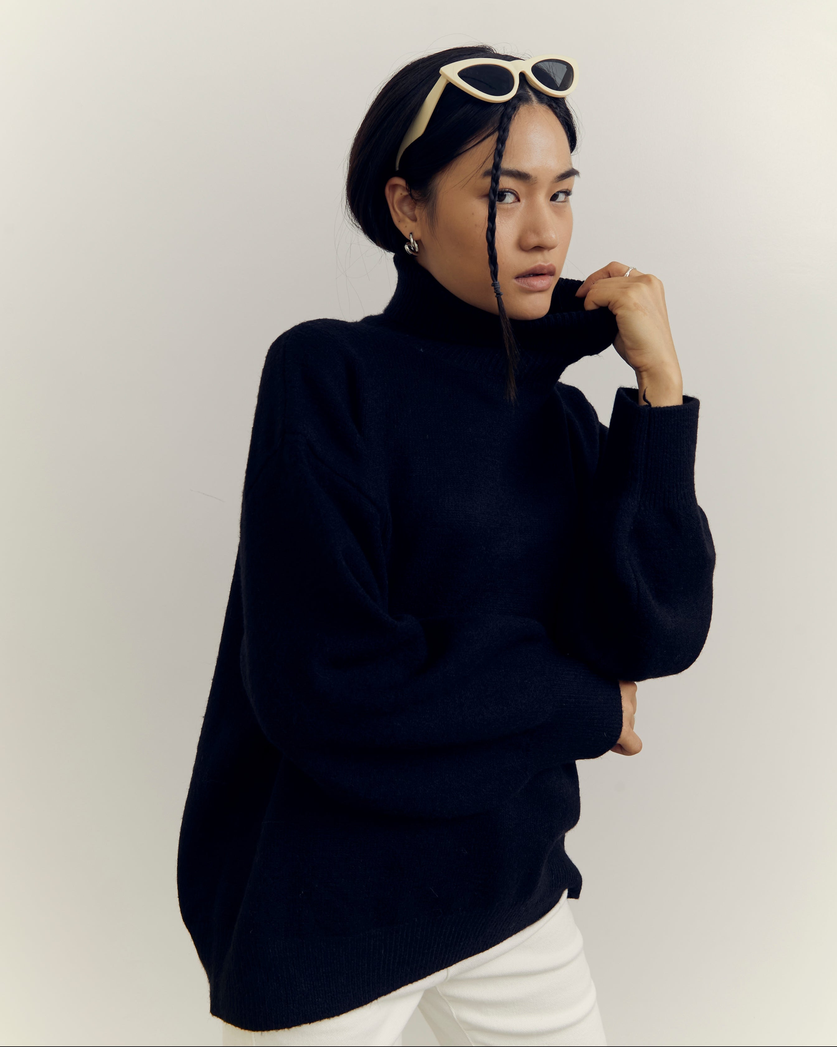 woman wearing a black turtleneck sweater and white pants against a plain background