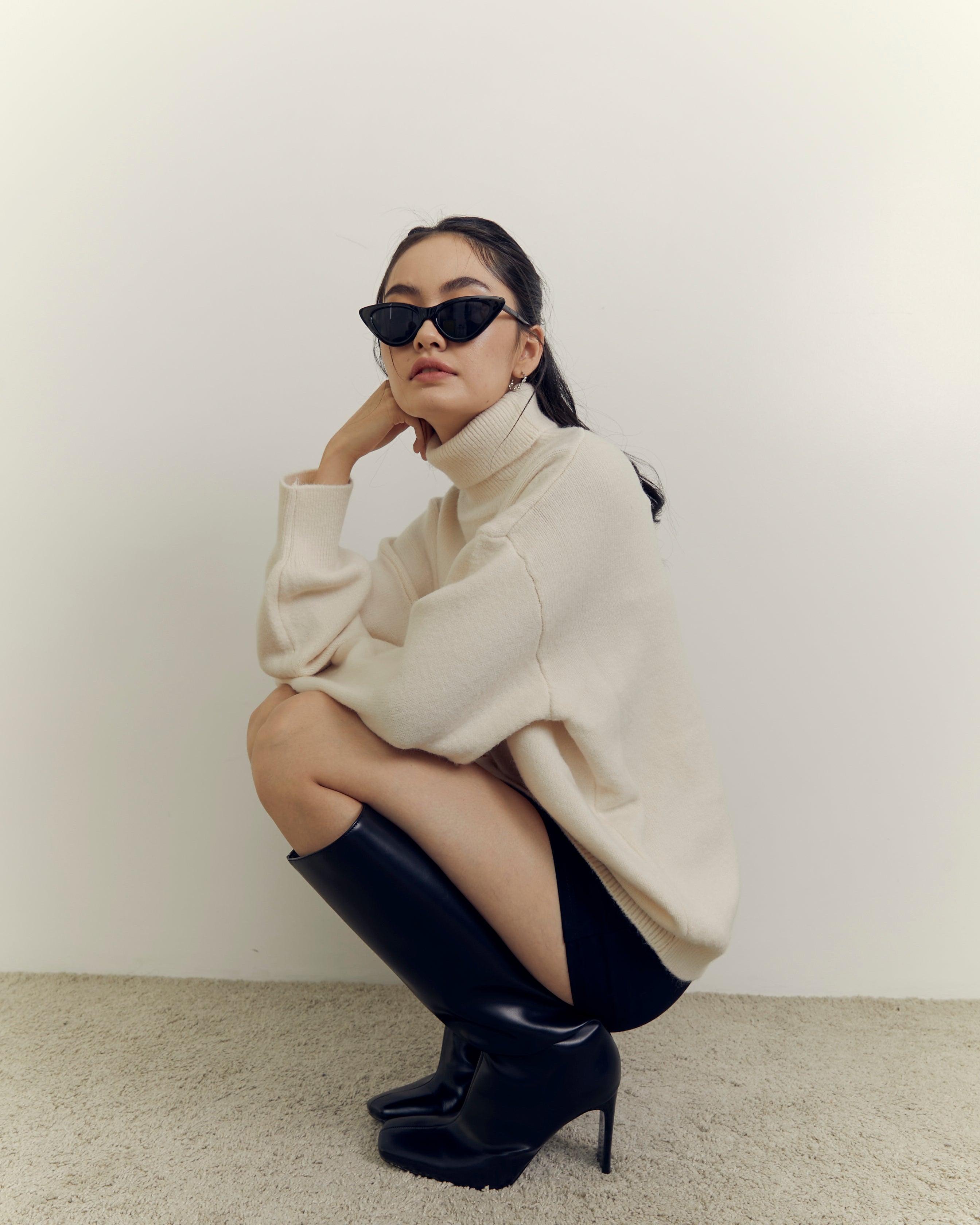 woman wearing a beige sweater and black boots sitting on the floor against a white wall.