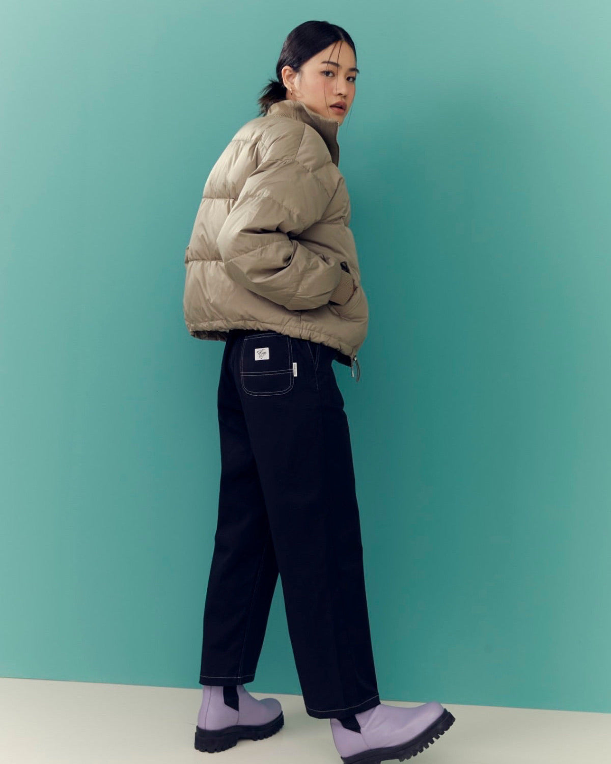 woman wearing a beige puffer jacket and dark pants against a teal background