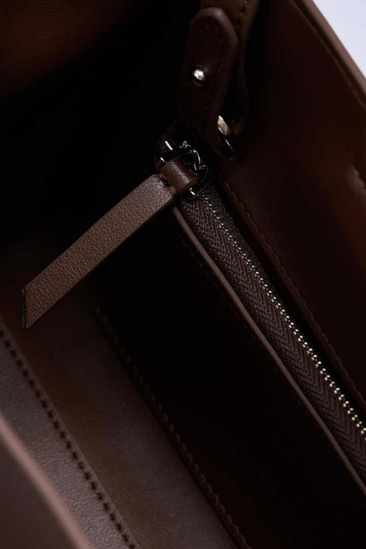 Close-up detail of a dark brown leather handbag showing a zipper with a pull tab and metal hardware.