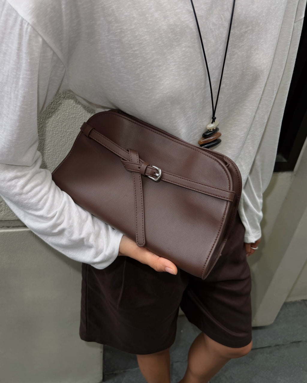 woman wearing a white long-sleeve top and dark brown shorts holds a brown leather crossbody bag with a matching strap and silver buckle.