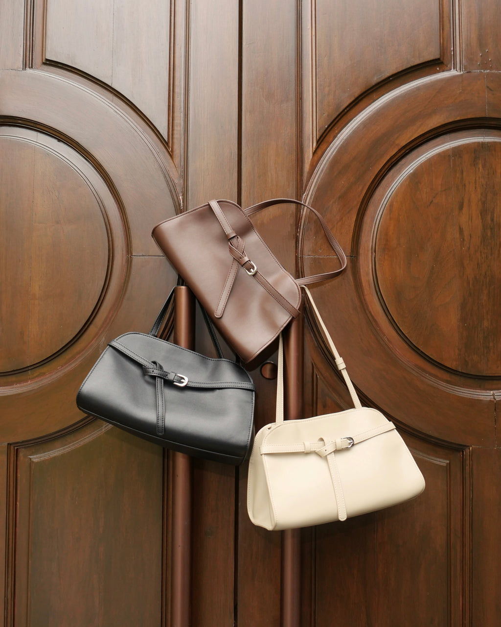 Three leather handbags in black, taupe-brown, and cream hang against a large dark wood paneled door.