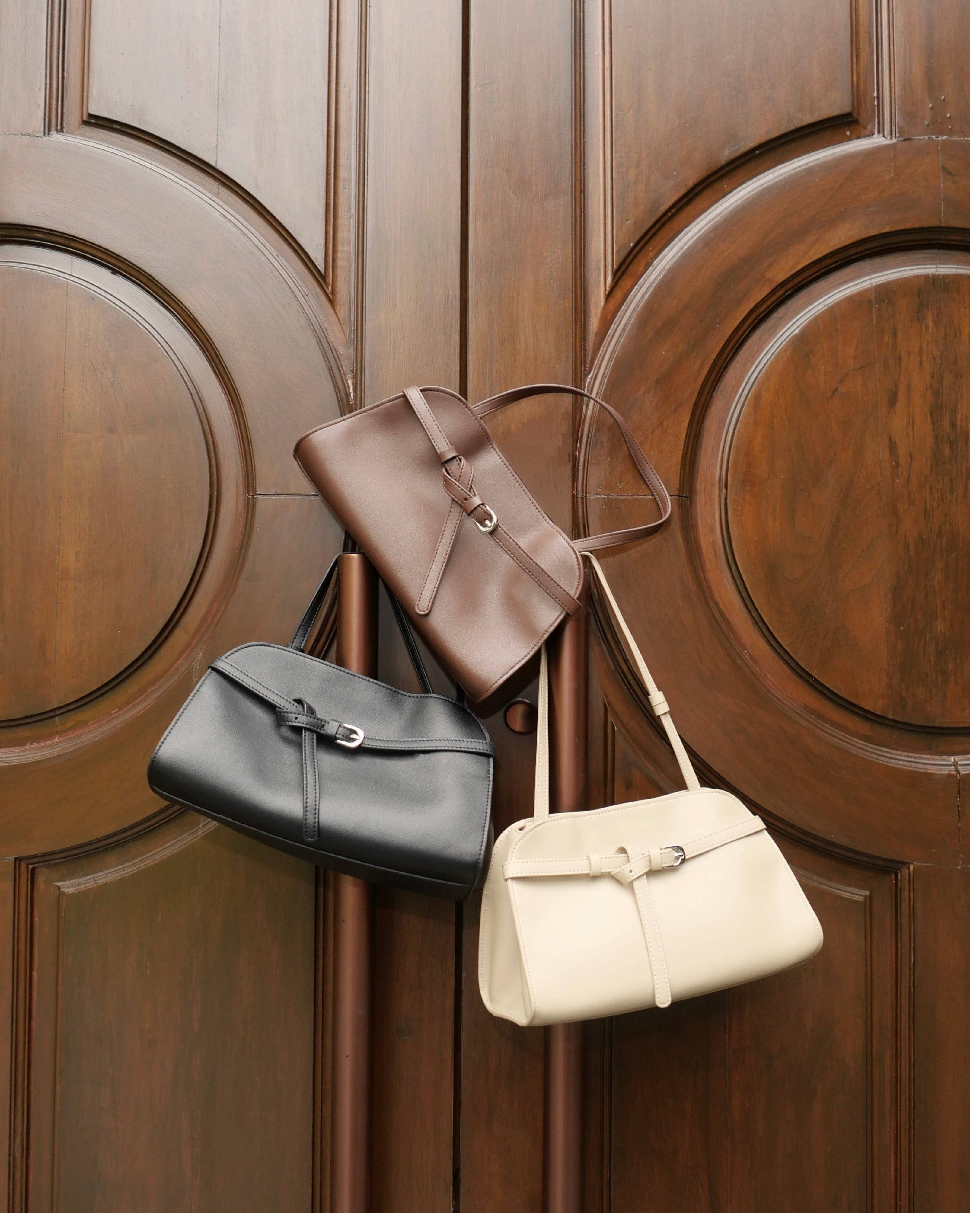 Three leather handbags in black, taupe-brown, and cream hang against a large dark wood paneled door.