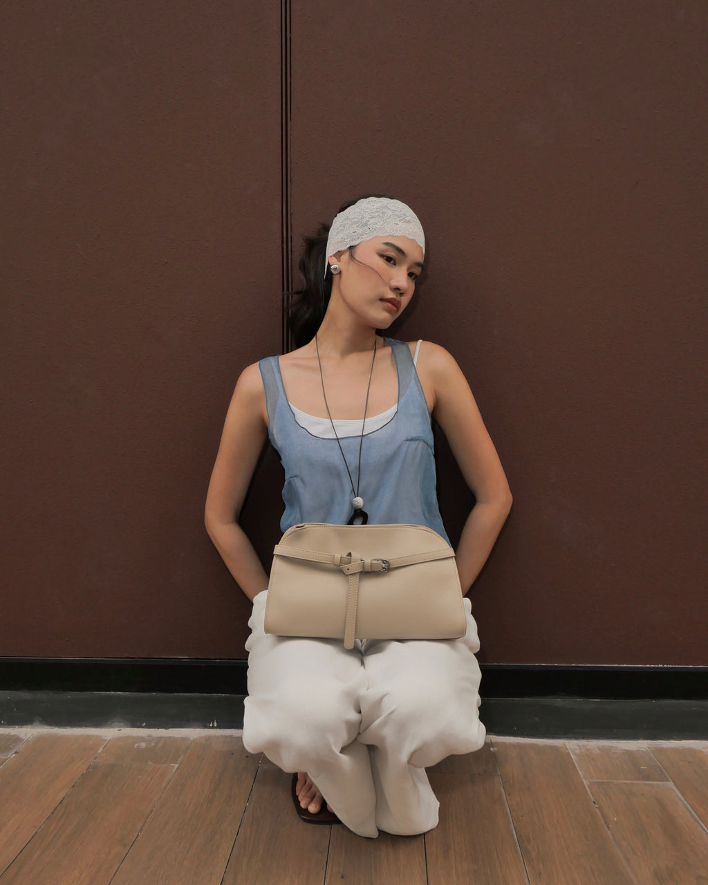 woman wearing a light blue sleeveless top, white pants, and a white headscarf sits against a dark brown wall, holding a beige shoulder bag.