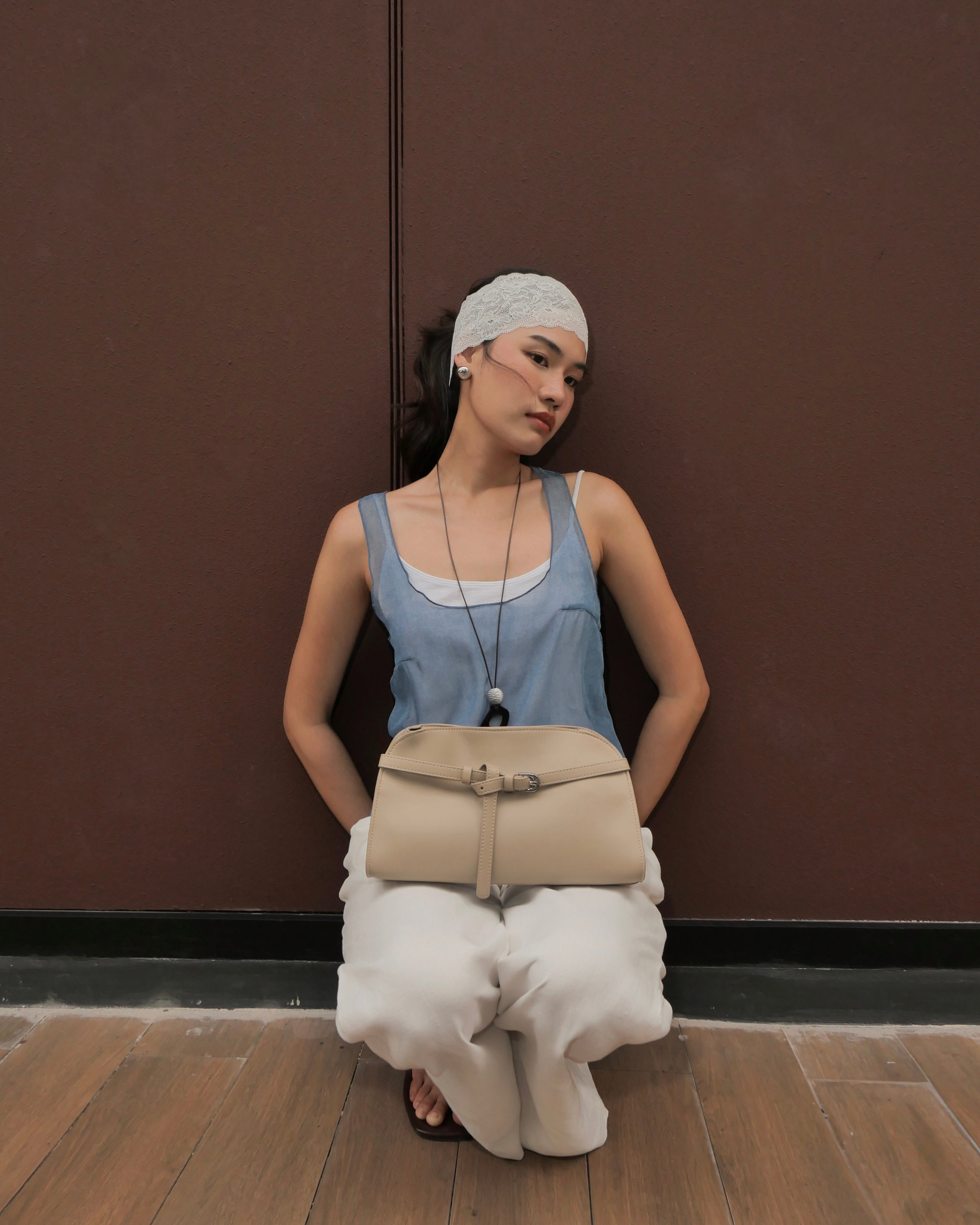 woman wearing a light blue sleeveless top, white pants, and a white headscarf sits against a dark brown wall, holding a beige shoulder bag.