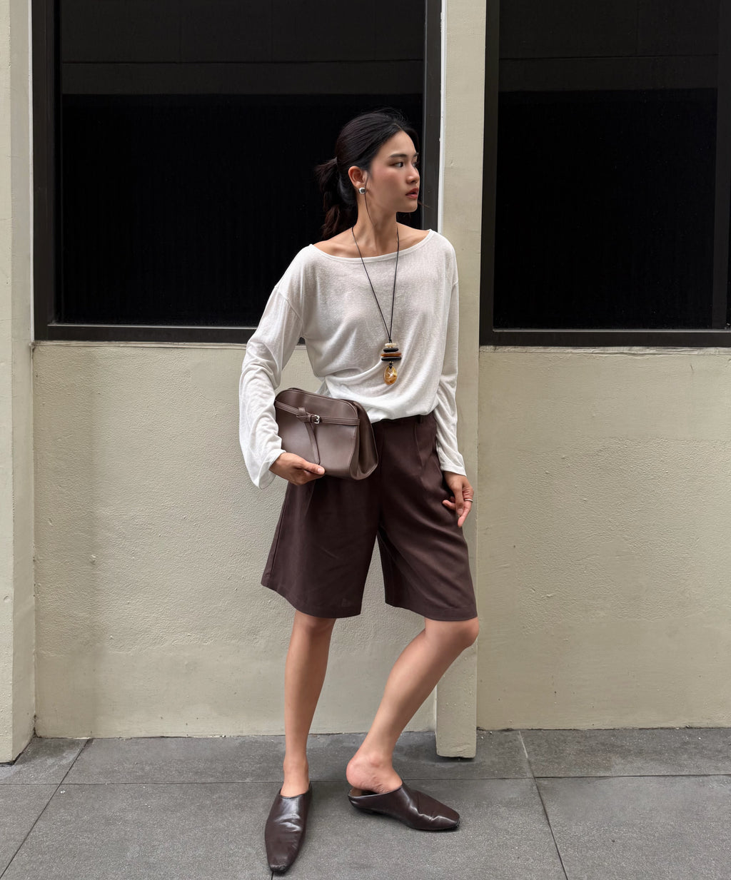 woman stands outdoors against a beige wall with dark window frames, wearing a white long-sleeve boat-neck top, dark brown shorts, dark brown mule loafers, and holding a taupe-brown leather crossbody bag.