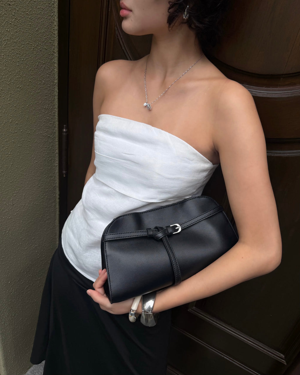 woman wearing a white strapless top and black skirt holds a black leather clutch bag with silver hardware, standing in front of a dark wooden door.