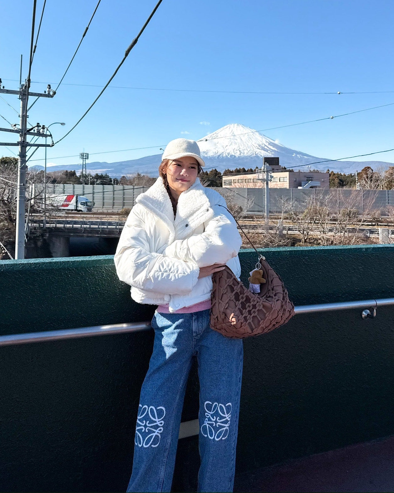 woman in a white coat and blue jeans standing on a bridge with Mount Fuji in the background