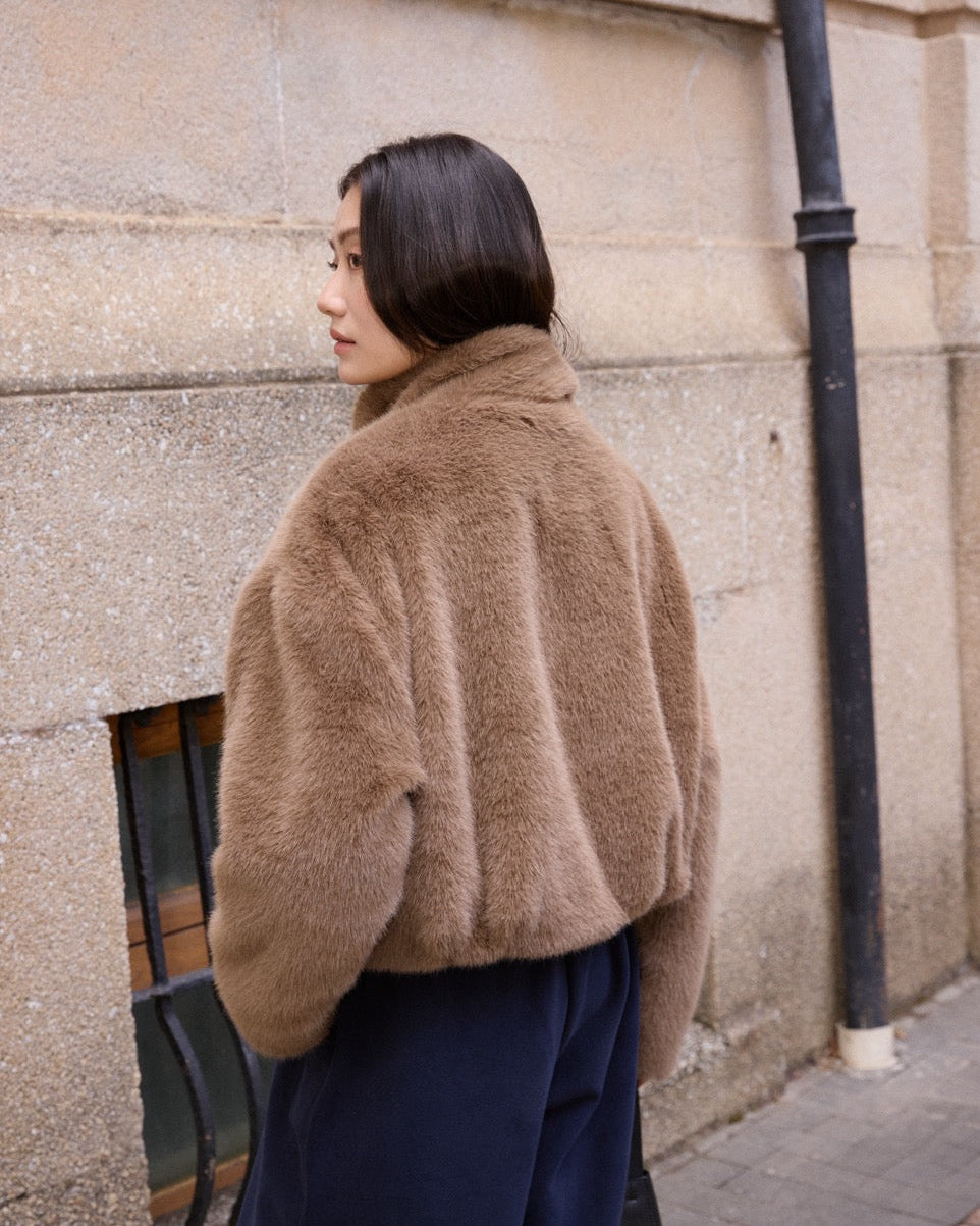 woman wearing a brown fuzzy jacket standing against a stone wall.