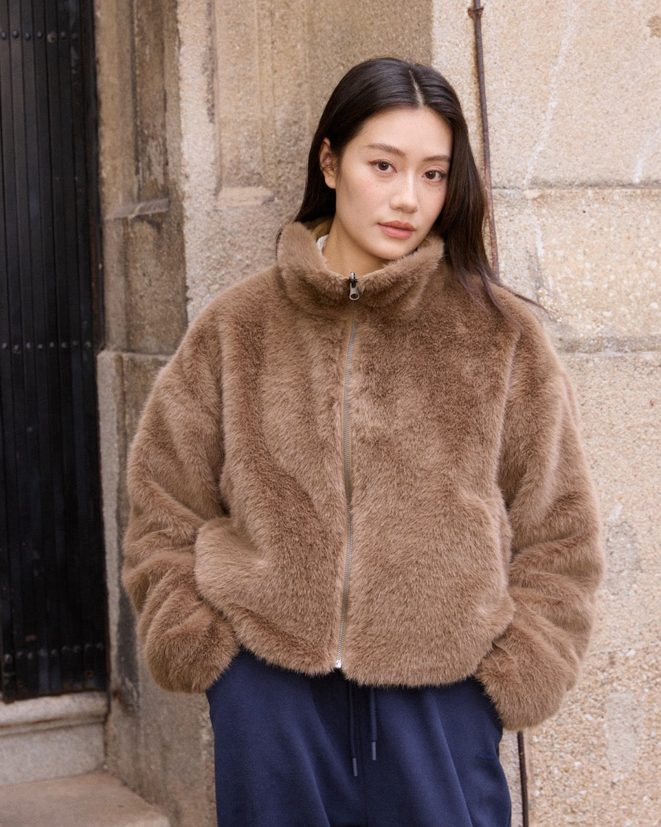 Woman wearing a brown fluffy jacket standing against a stone wall.