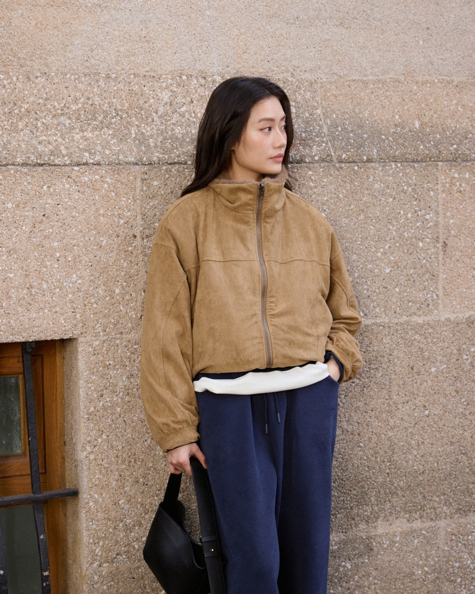 Woman wearing a tan jacket and blue pants against a textured wall.