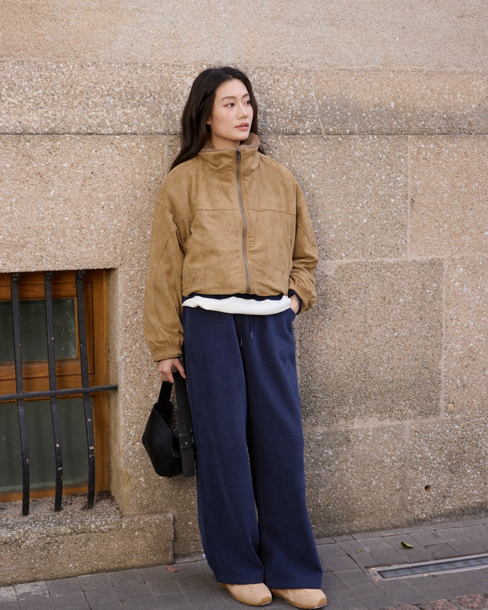 Woman wearing a beige jacket and blue pants standing against a textured wall.