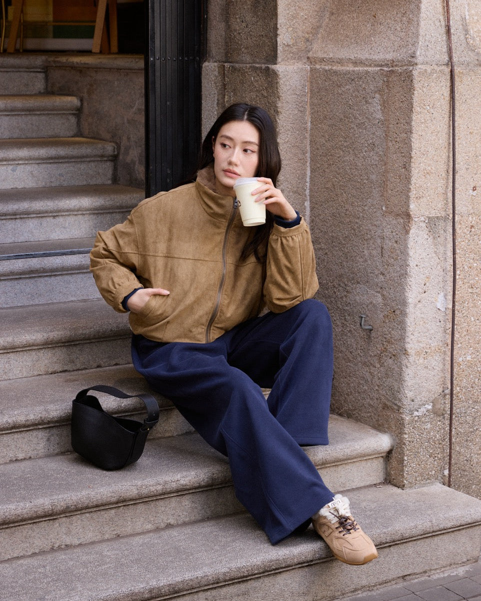 Woman sitting on steps holding a coffee cup, wearing a brown jacket and blue pants.