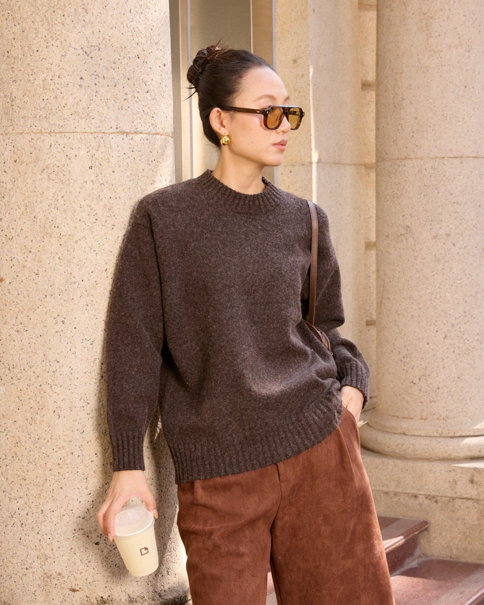 Woman wearing a dark brown sweater and brown pants standing against a stone wall.