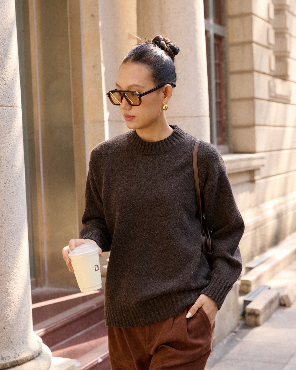 Woman wearing a dark sweater and brown pants, holding a coffee cup, standing in front of a building.
