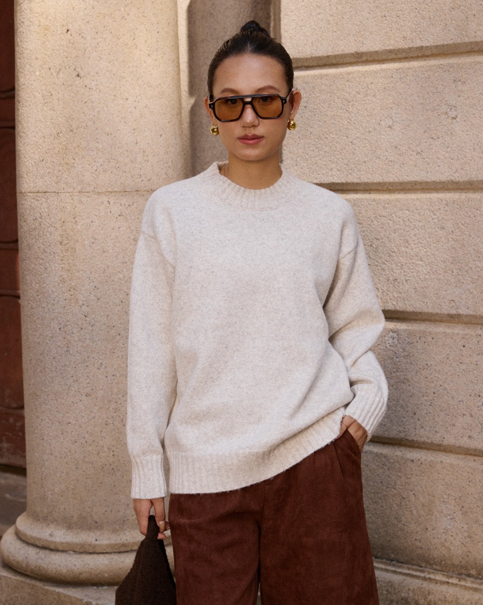 woman  wearing a light sweater and brown pants standing against a stone wall.