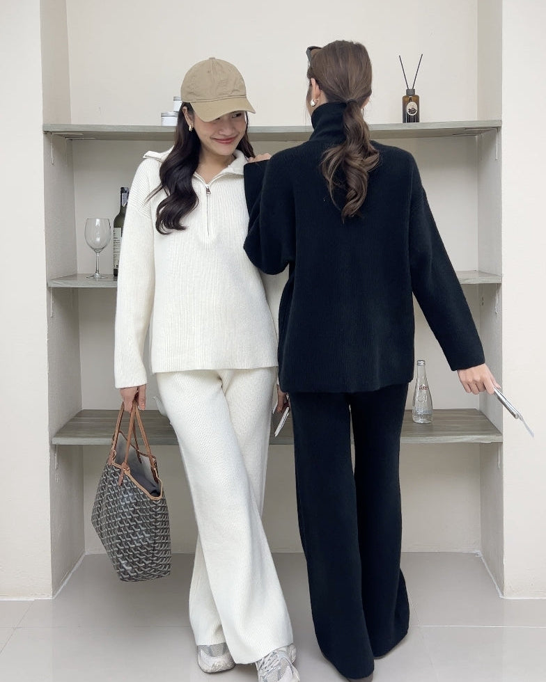 Two women, one in a white outfit and the other in black, standing in a minimalistic room.