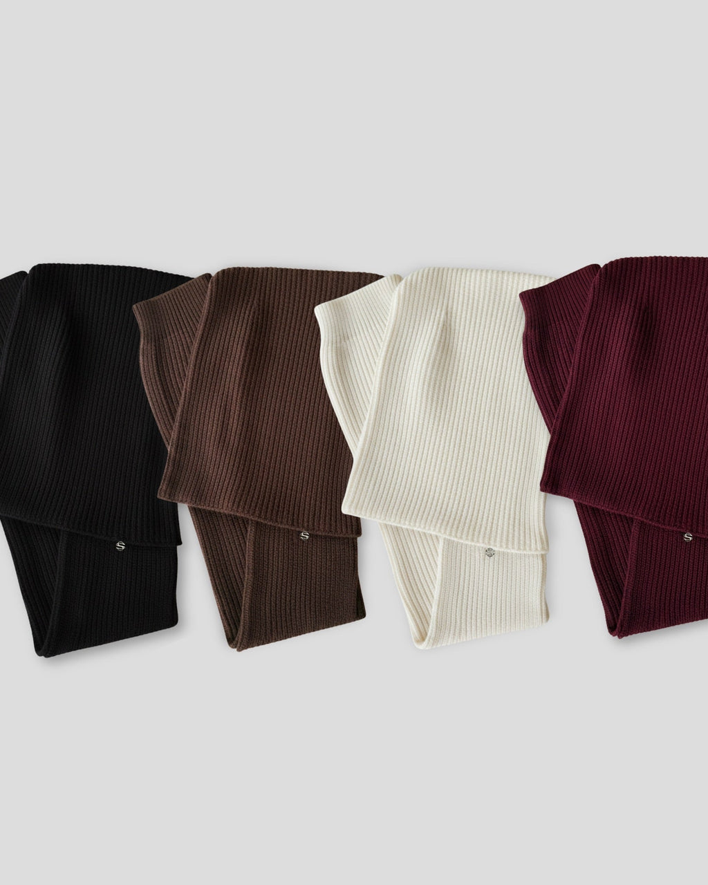Four folded ribbed sweaters in black, brown, white, and burgundy on a light gray background.