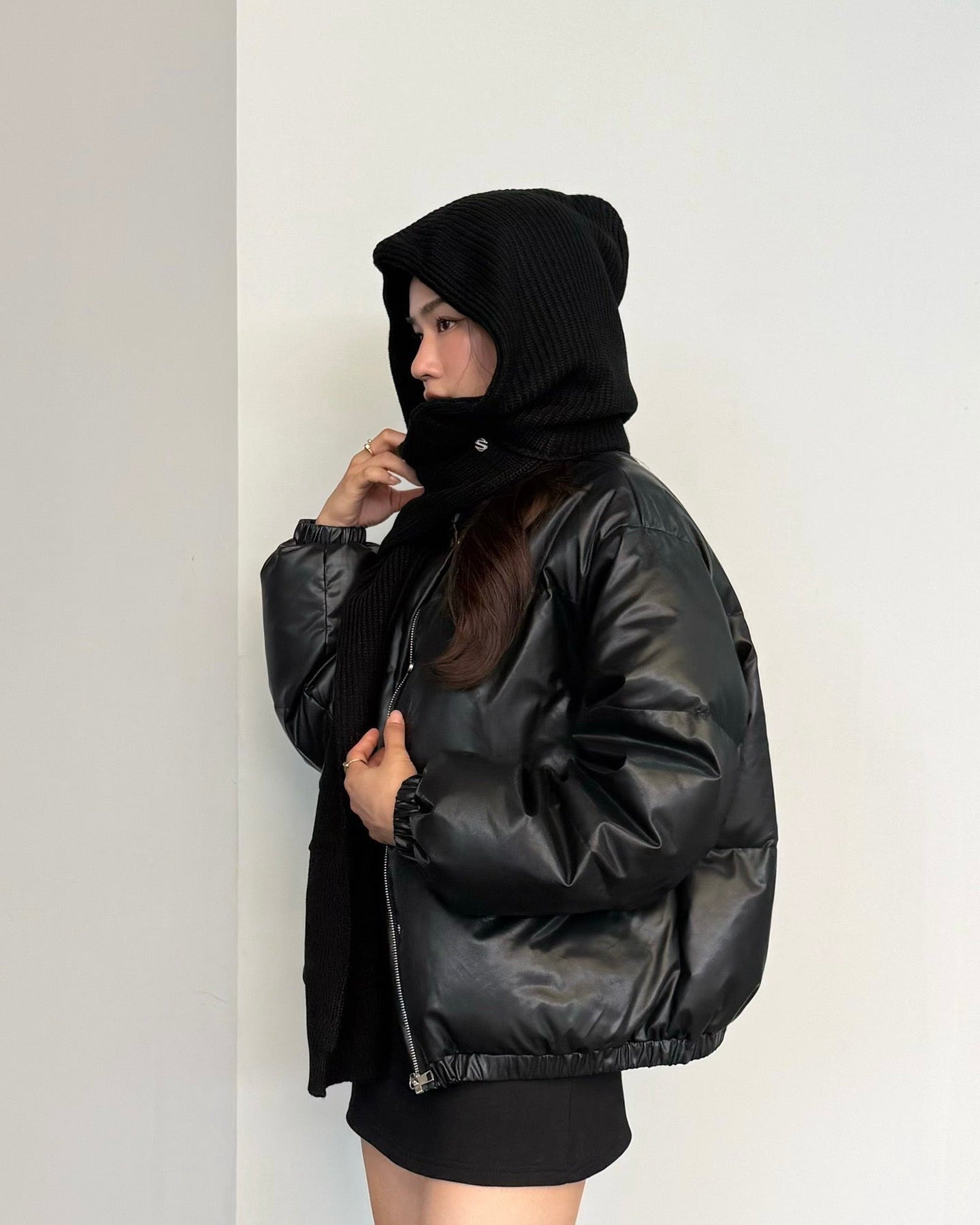 woman wearing a black puffer jacket with a hood against a white wall.