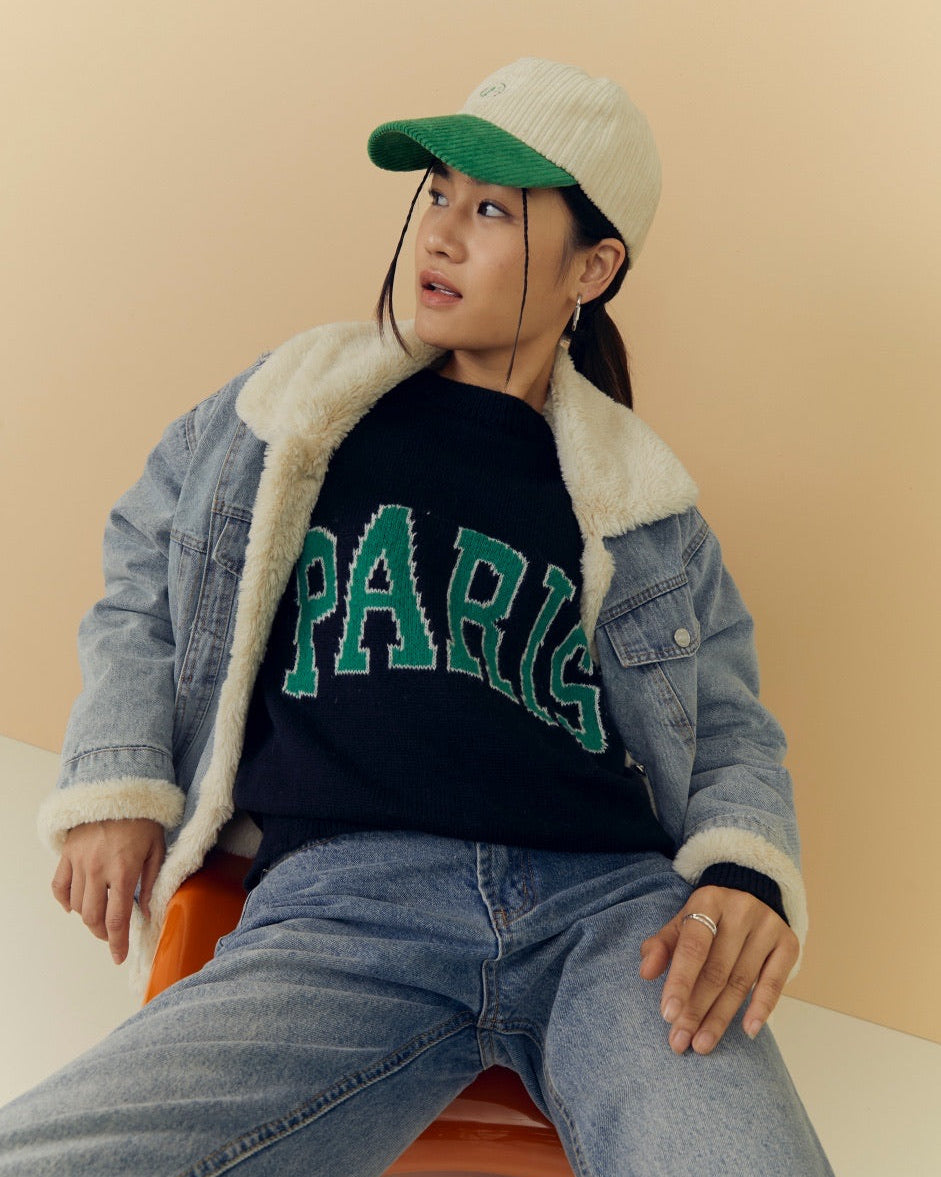 woman wearing a denim jacket with shearling lining, black sweater with 'PARIS' text, and green cap on a beige background