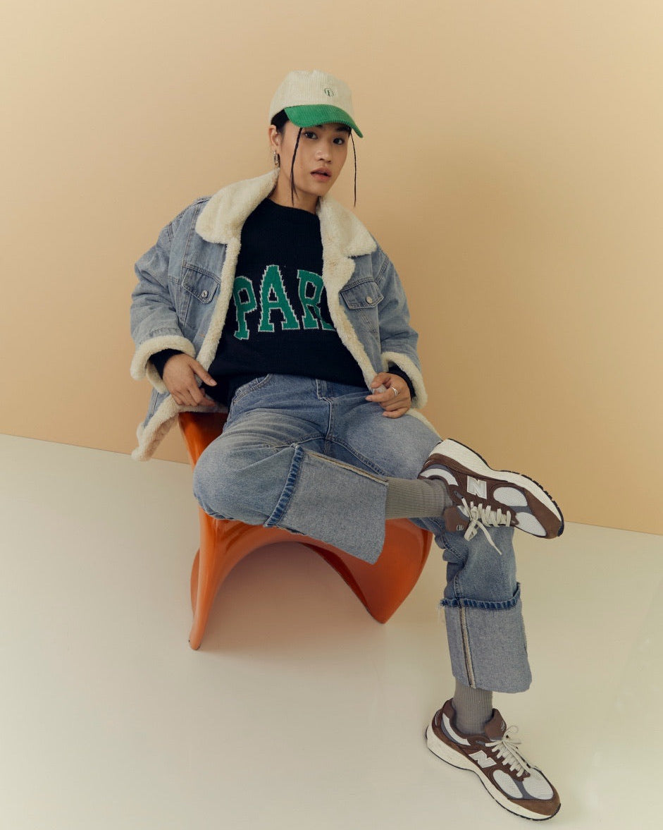 woman sitting on an orange chair wearing a denim jacket, black t-shirt with green text, blue jeans, and black sneakers.