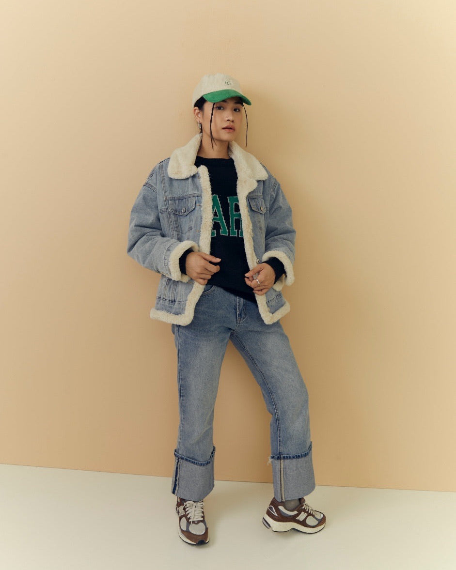 woman wearing a denim jacket with white fur lining, jeans, and a green cap against a beige background