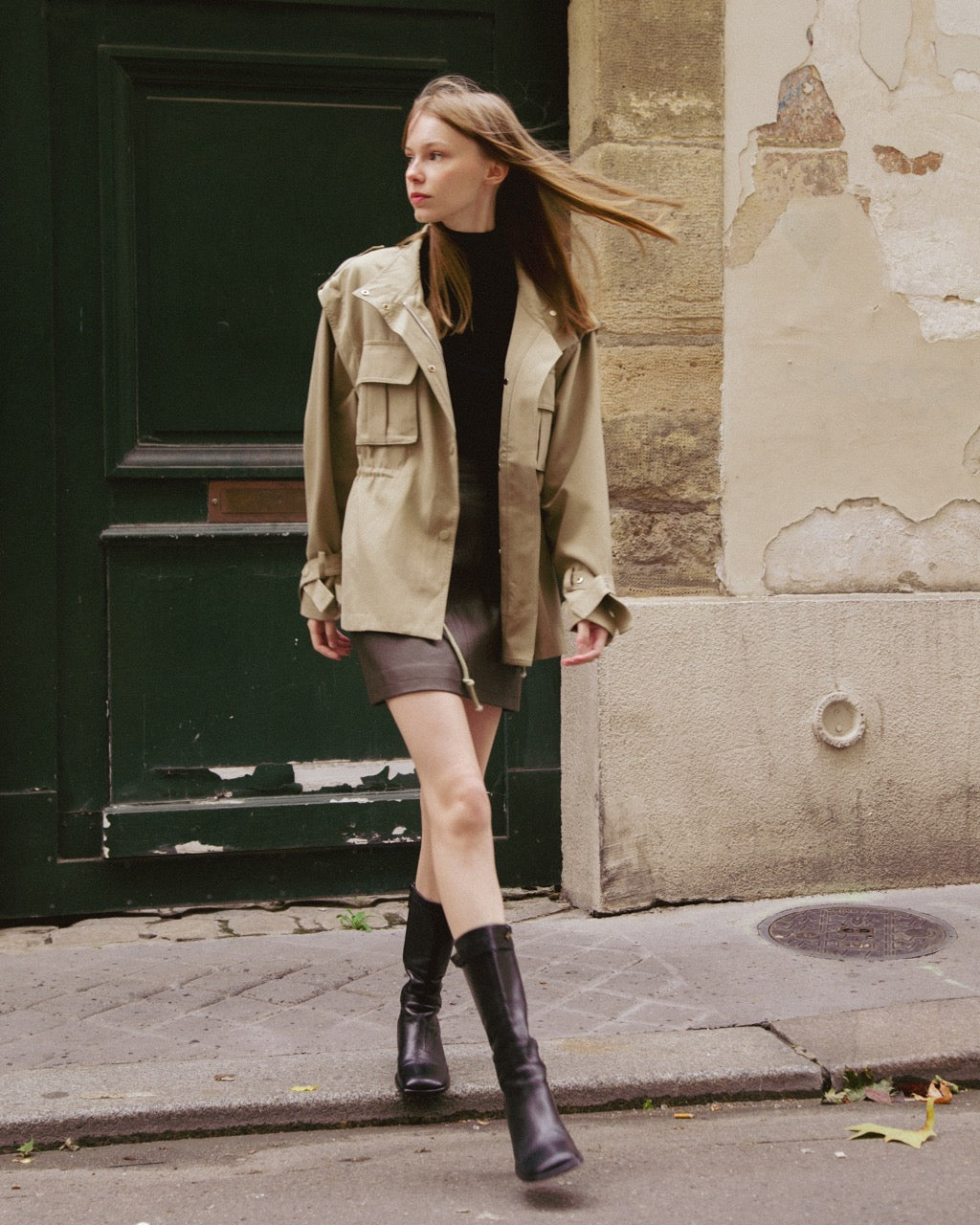 Woman in a olive green trench coat and black boots walking on a street.