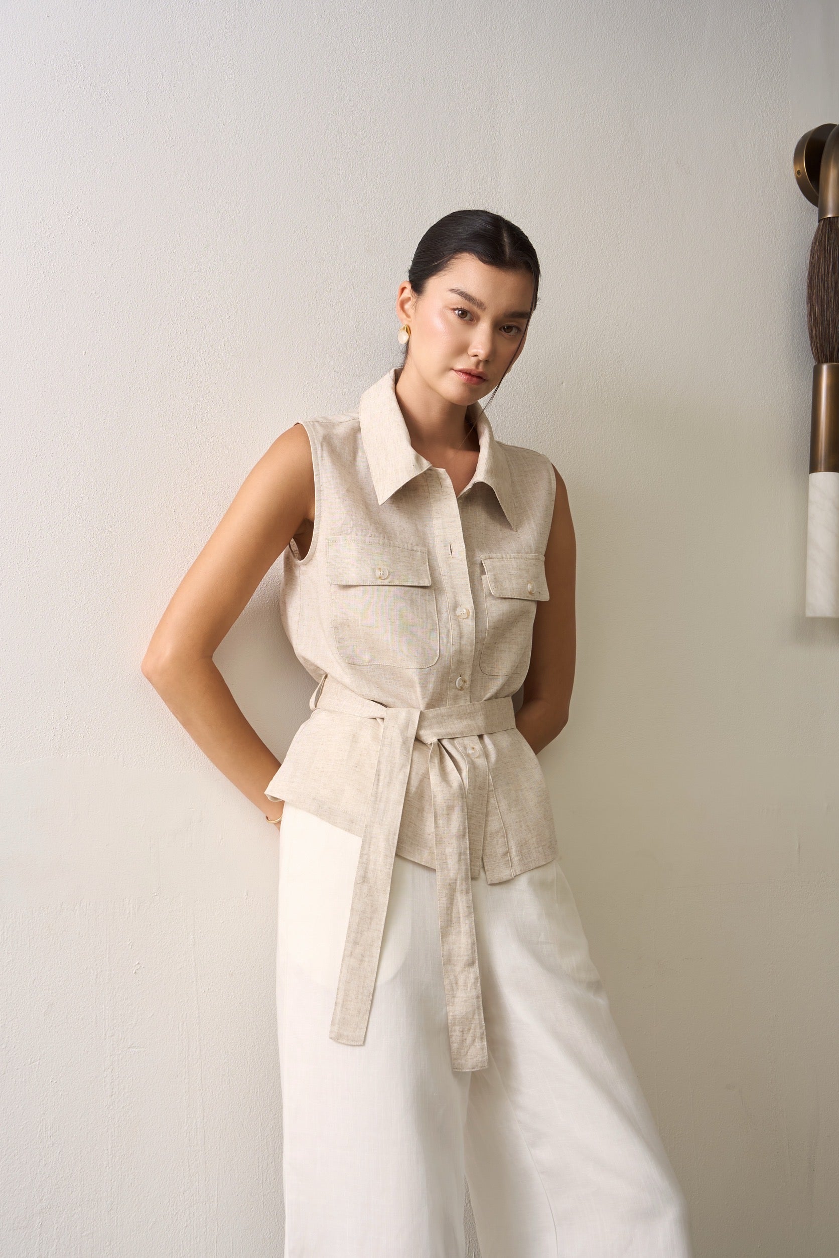 Woman wearing a beige sleeveless double pocket linen shirt with a belt and white linen pants against a plain background