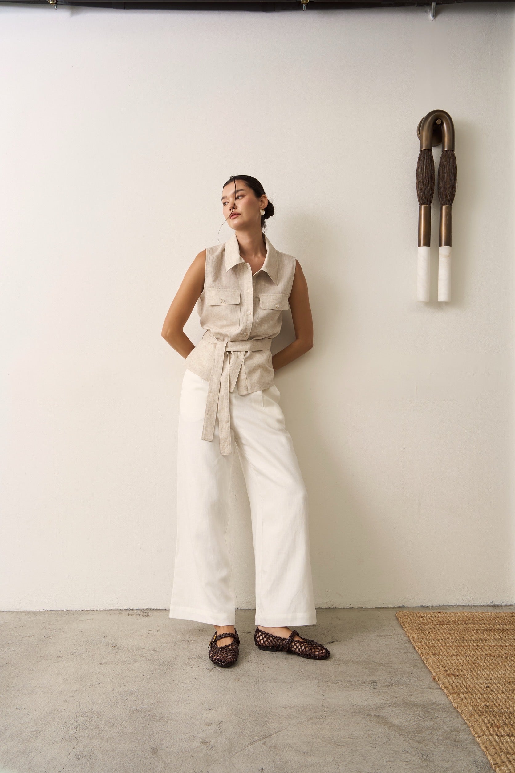 Woman in beige sleeveless linen top with belt and white linen trousers standing against a white wall with a decorative object.