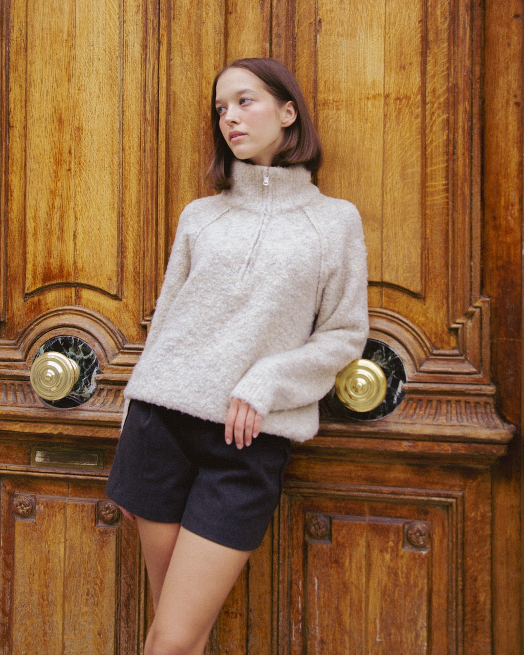 Woman wearing a light gray sweater and black shorts standing in front of a wooden door.