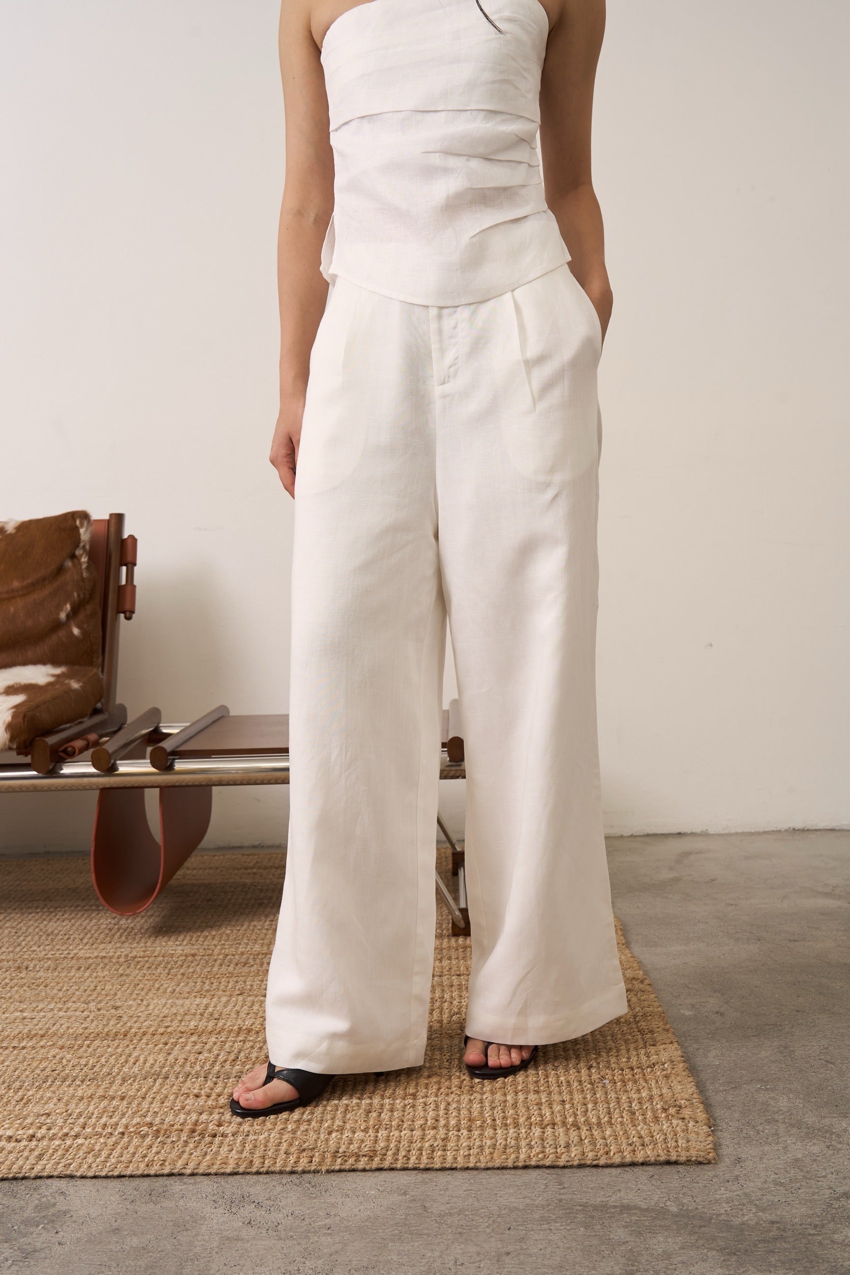 Close up of a woman wearing a white strapless linen top and wide-leg linen pants standing on a mat with a white wall and wooden chair in the background.