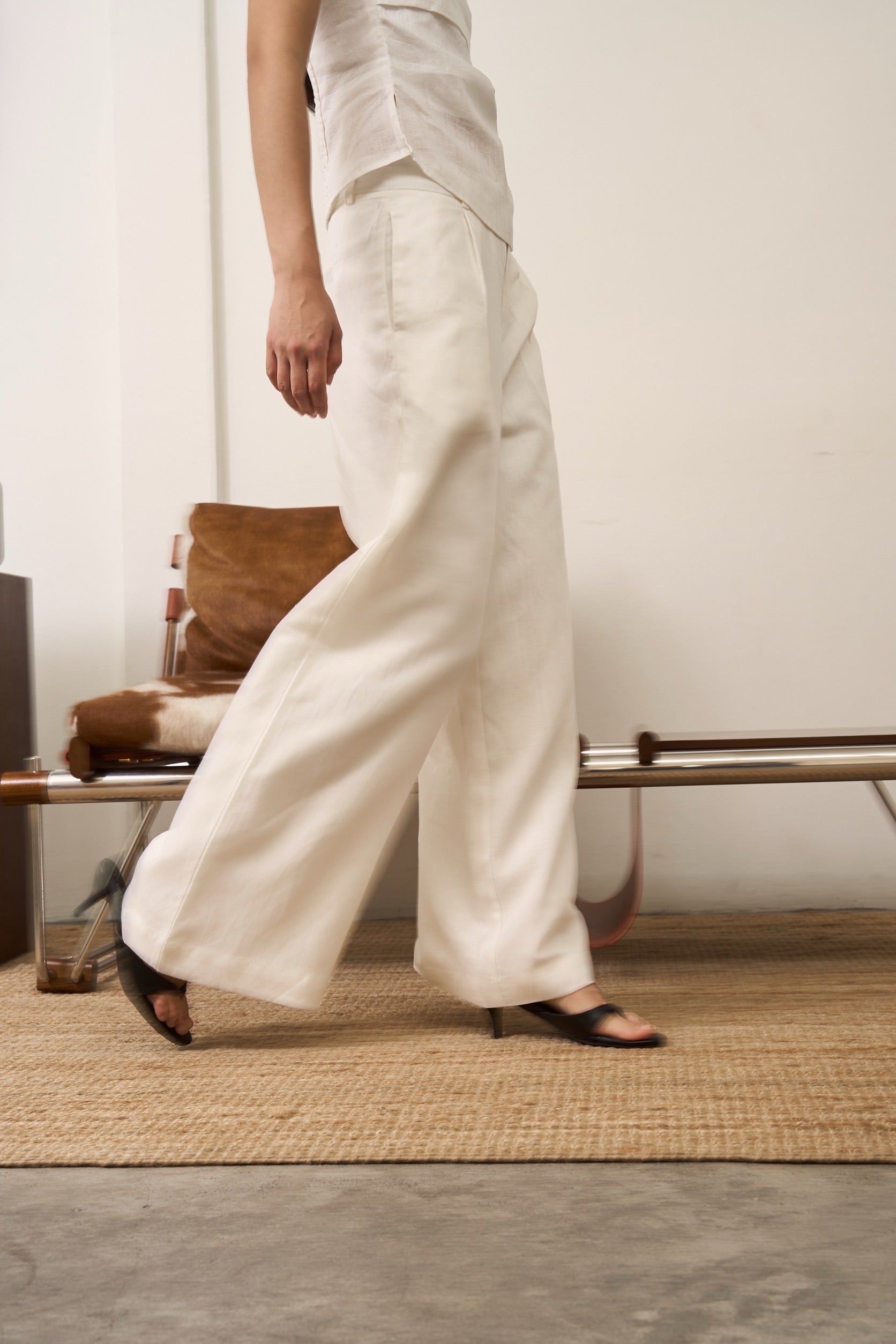 Close up of a woman wearing white linen sleeveless shirt and pants standing on a wooden floor with a chair and rug in the background.