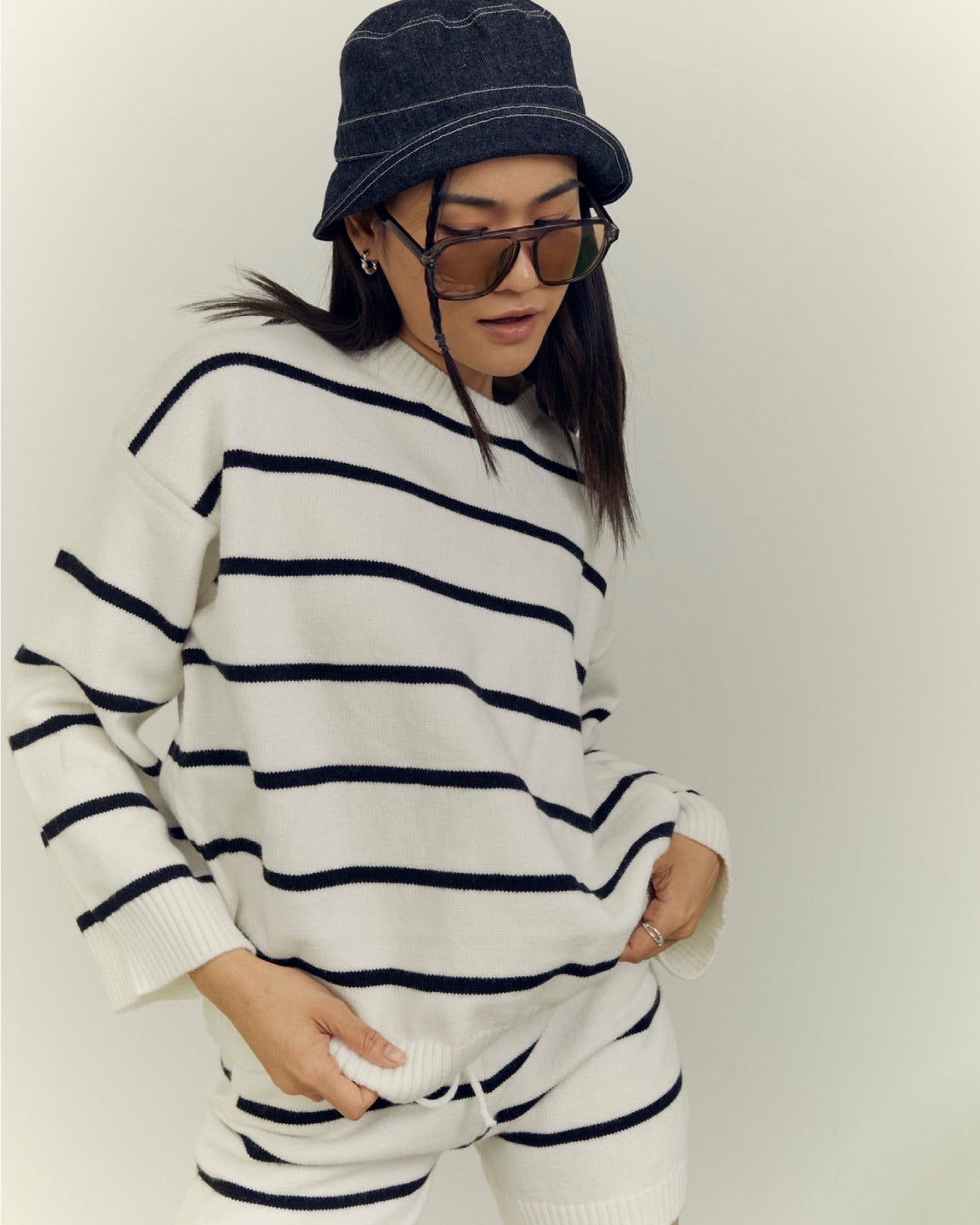 woman wearing a white and navy striped sweater with matching shorts, dark denim bucket hat, and sunglasses on a plain background.