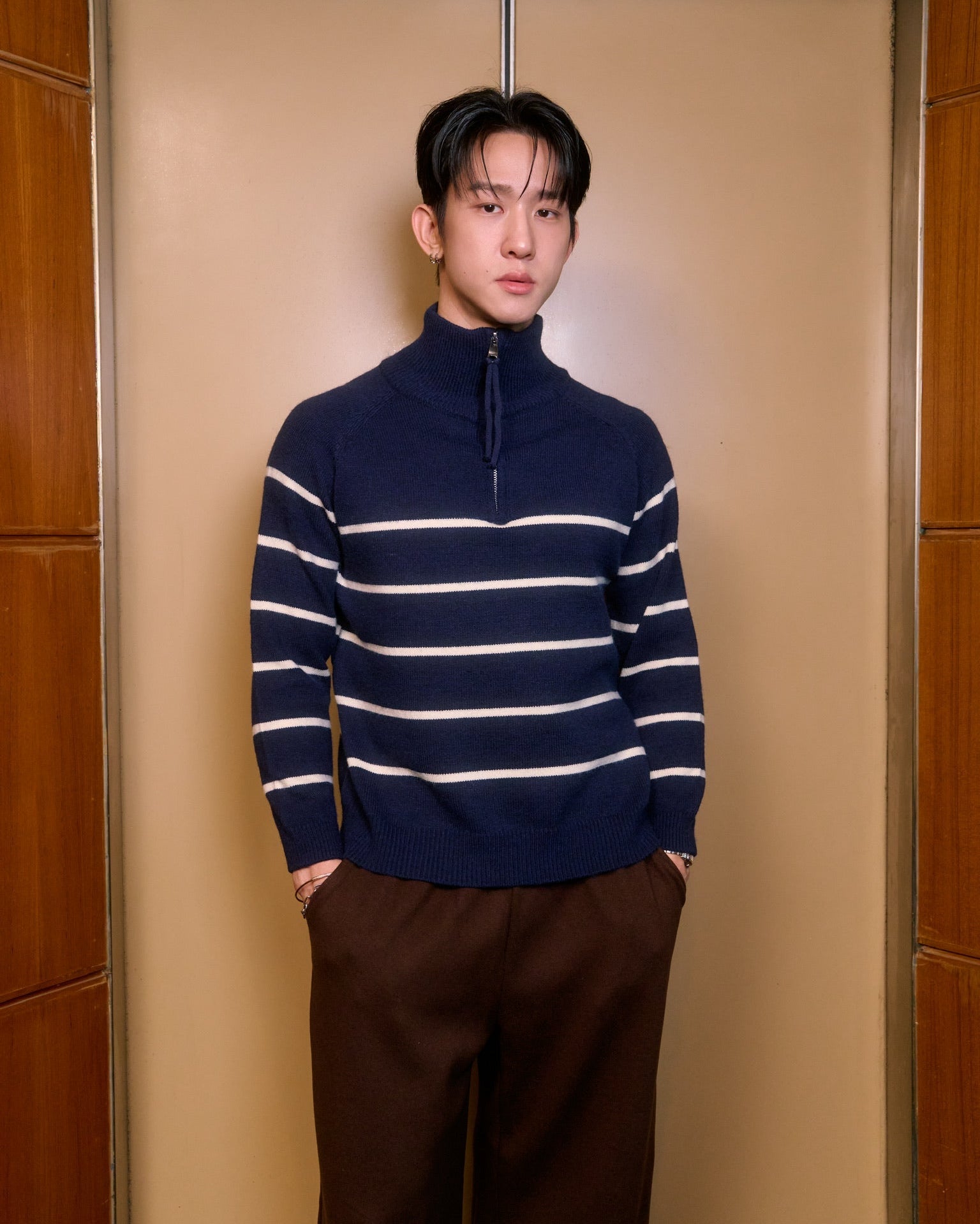 man wearing a navy blue and white striped sweater with brown pants standing against a beige wall.