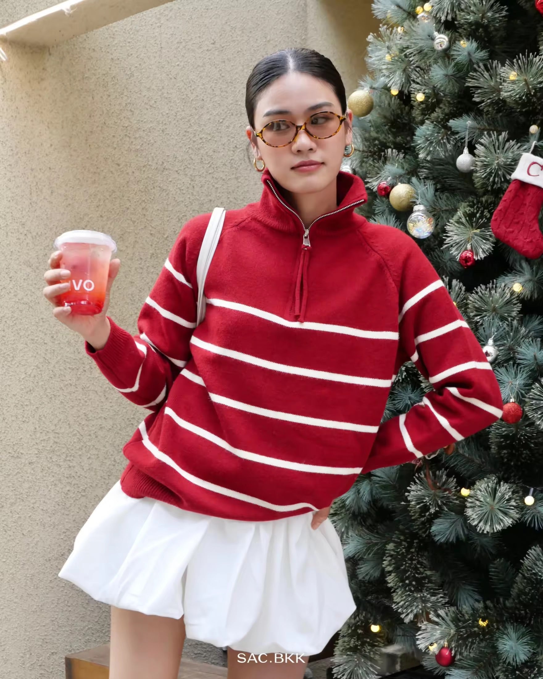 woman wearing a red and white striped sweater holding a drink, standing next to a decorated Christmas tree.