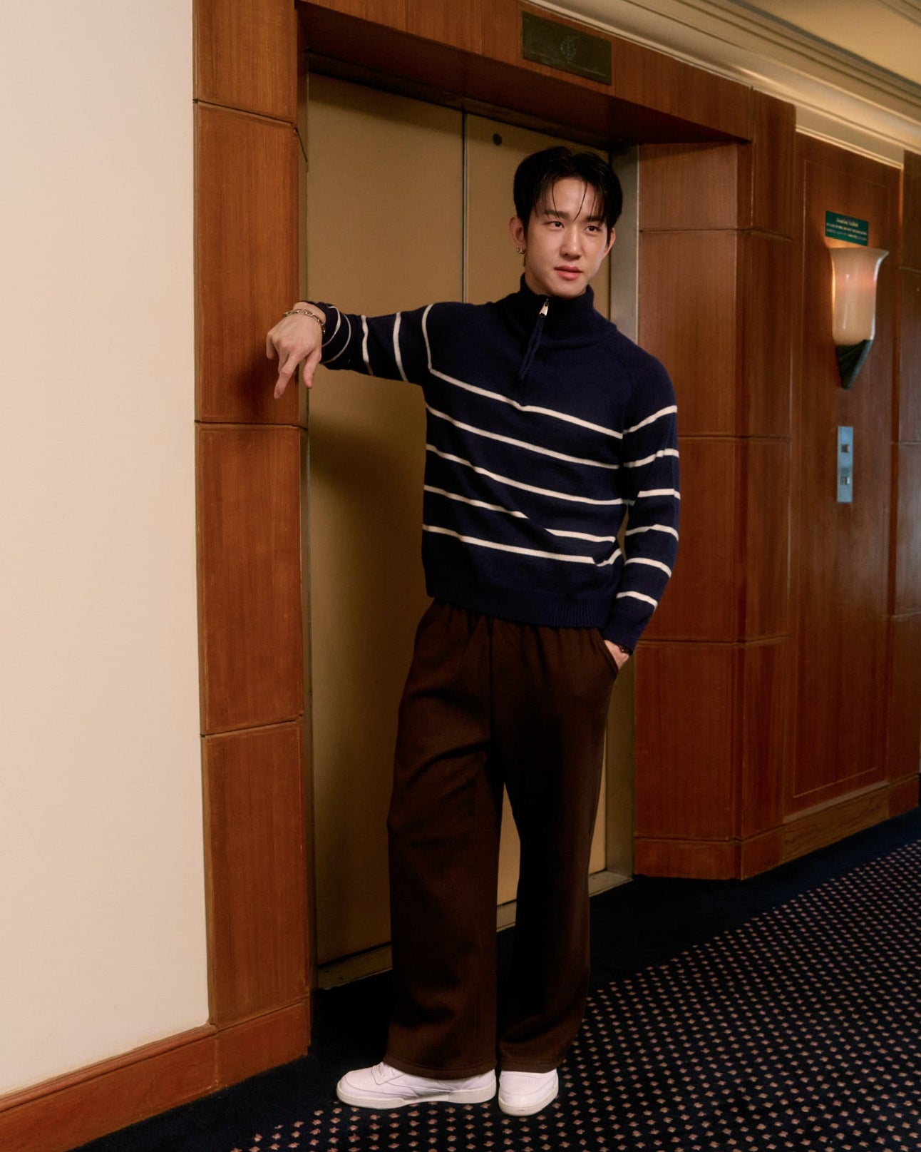 Man standing in a hallway with wooden doors and patterned floor.